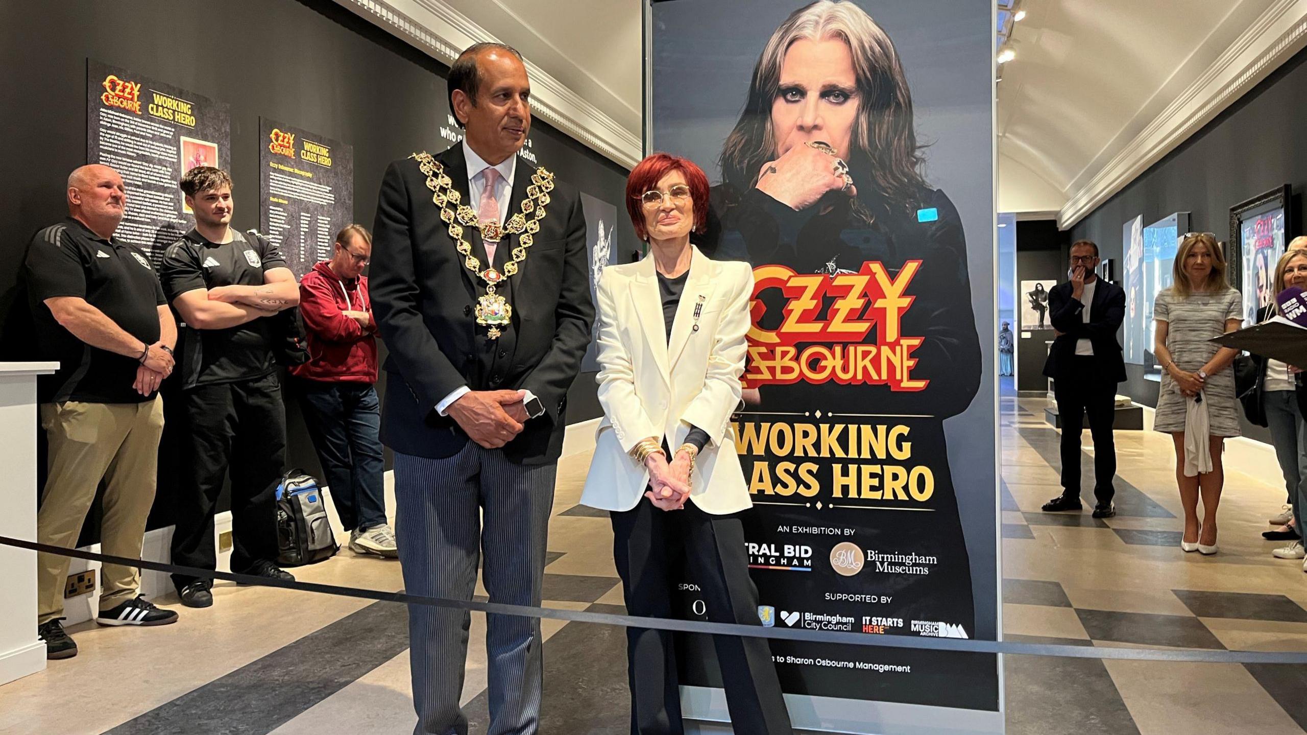 A man and woman stand in an exhibition hall, in front of a poster of a man that says "Ozzy Osbourne, Working Class Hero" on it and a black ribbon. The man is tall and wears grey suit trousers, a black suit jacket, white shirt, pale pink tie and a mayoral chain. The woman has dyed red hair, glasses and is wearing a white suit jacket with a black top underneath and black trousers. There are people standing around the edges of the exhibition hall looking on.