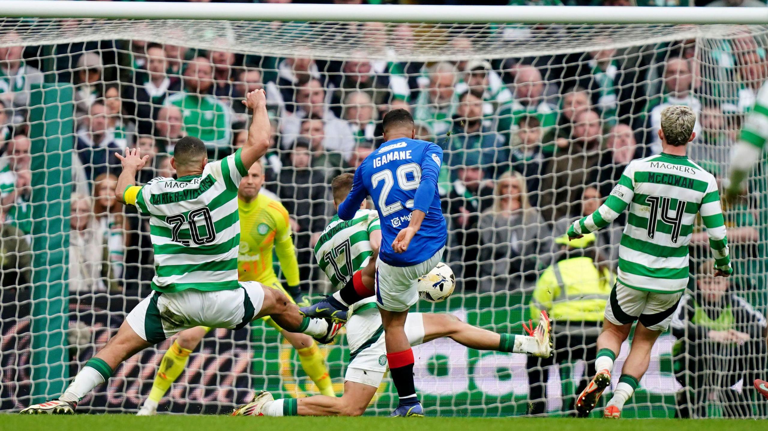 A footballer wearing a blue shift, white shorts and black and red socks kicks a ball towards a goalkeeper's top left-hand corner as three players, wearing green and white hoops fail to stop him.