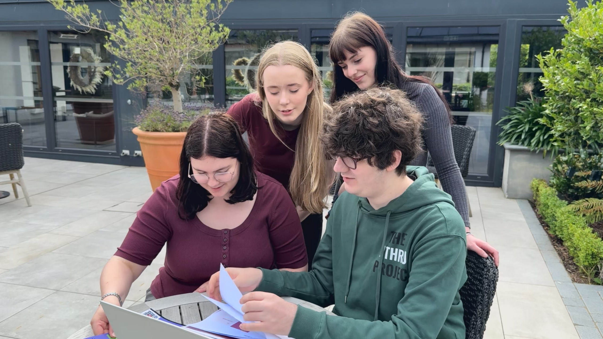 Four young people, two standing and two sitting, are looking at a laptop and some paper.