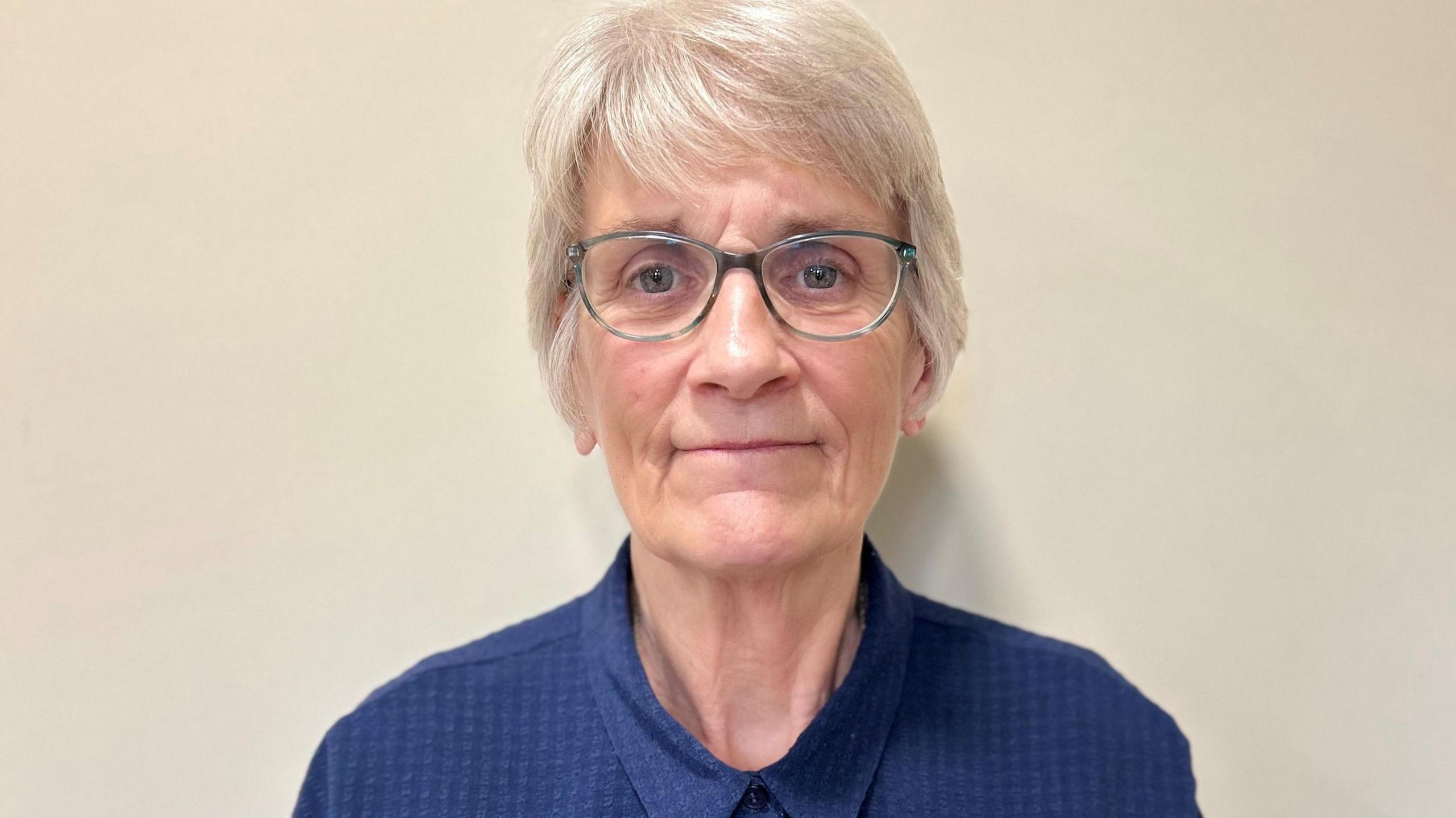 Angela Stone standing against a cream-painted wall. She has short silvery hair and is wearing grey-framed glasses and a blue shirt done up at the neck. She is looking towards the camera