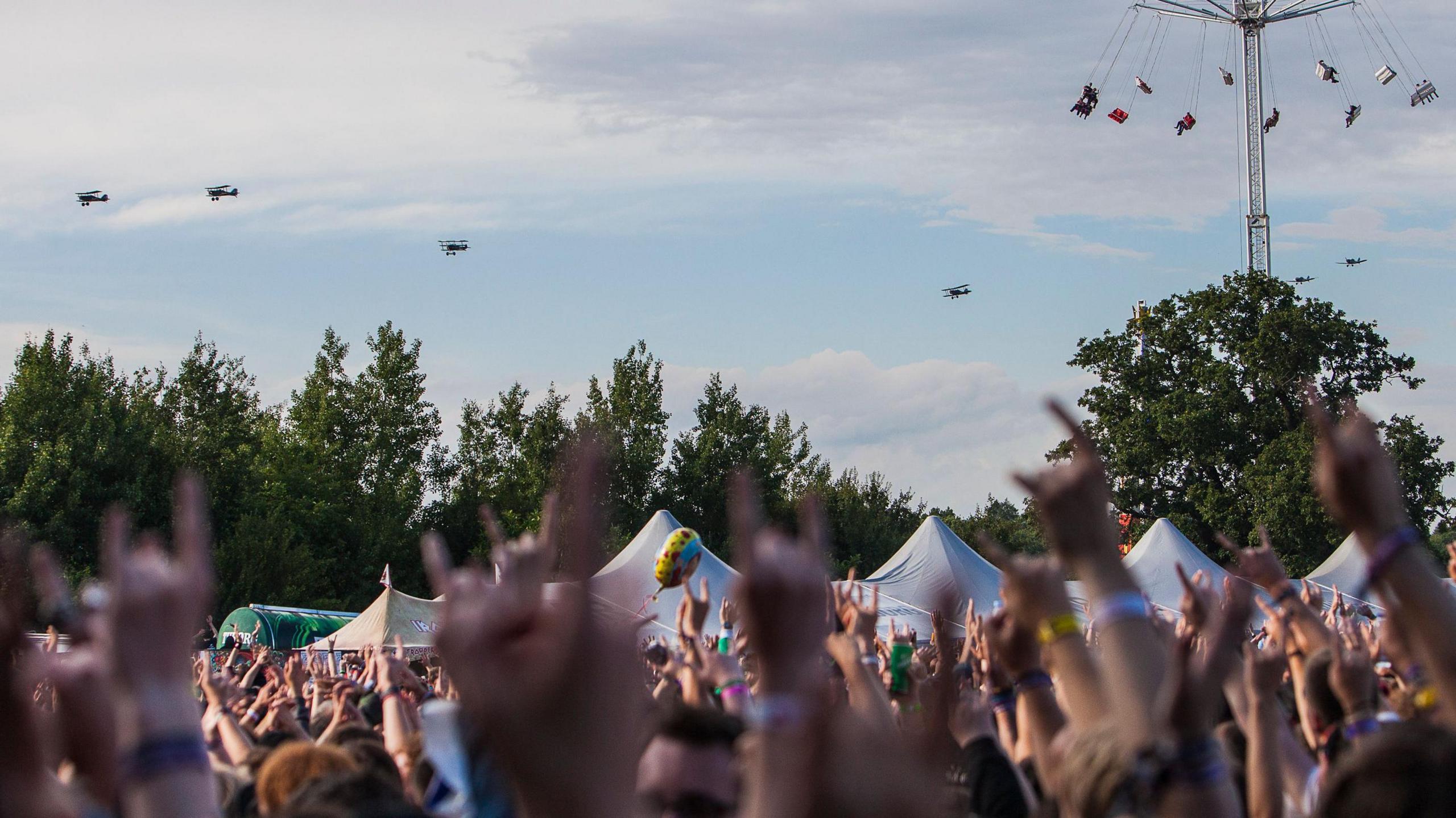 Crowd at an outdoor festival raising hands with 'rock on' gestures while World War One planes fly in the sky above.