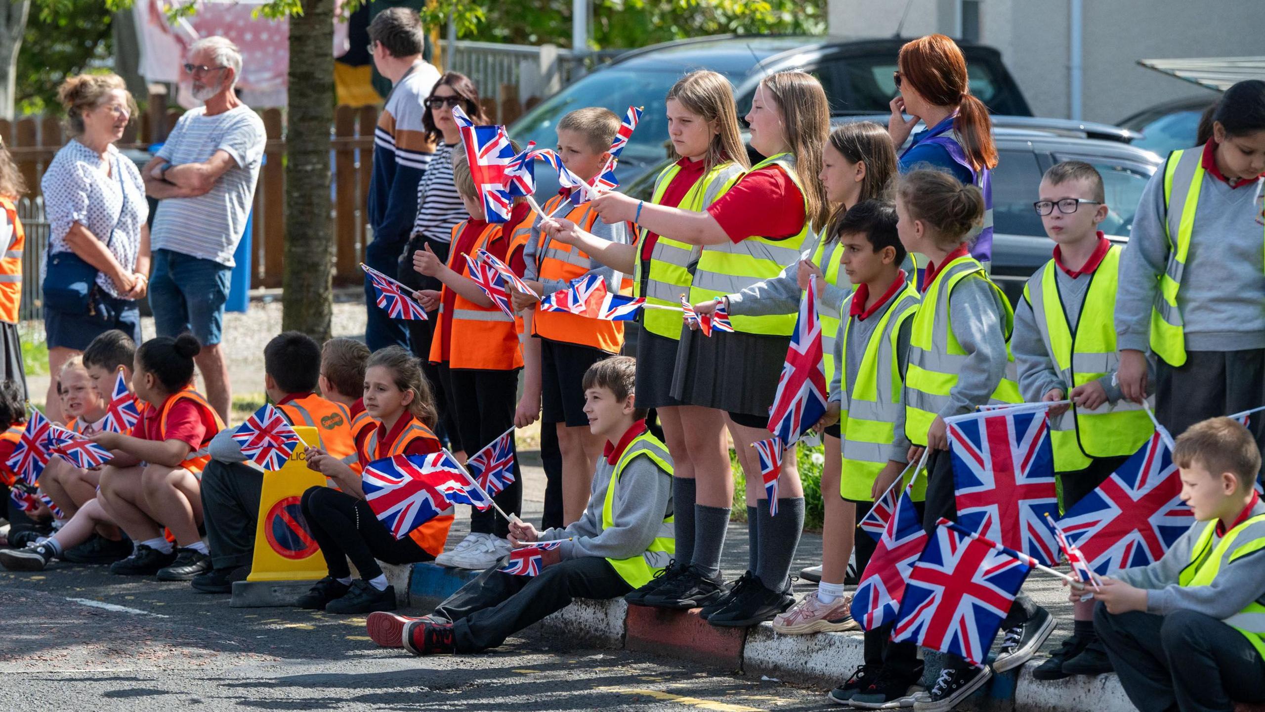 A group of young children sit and stand on a kerbside, holding union flags.