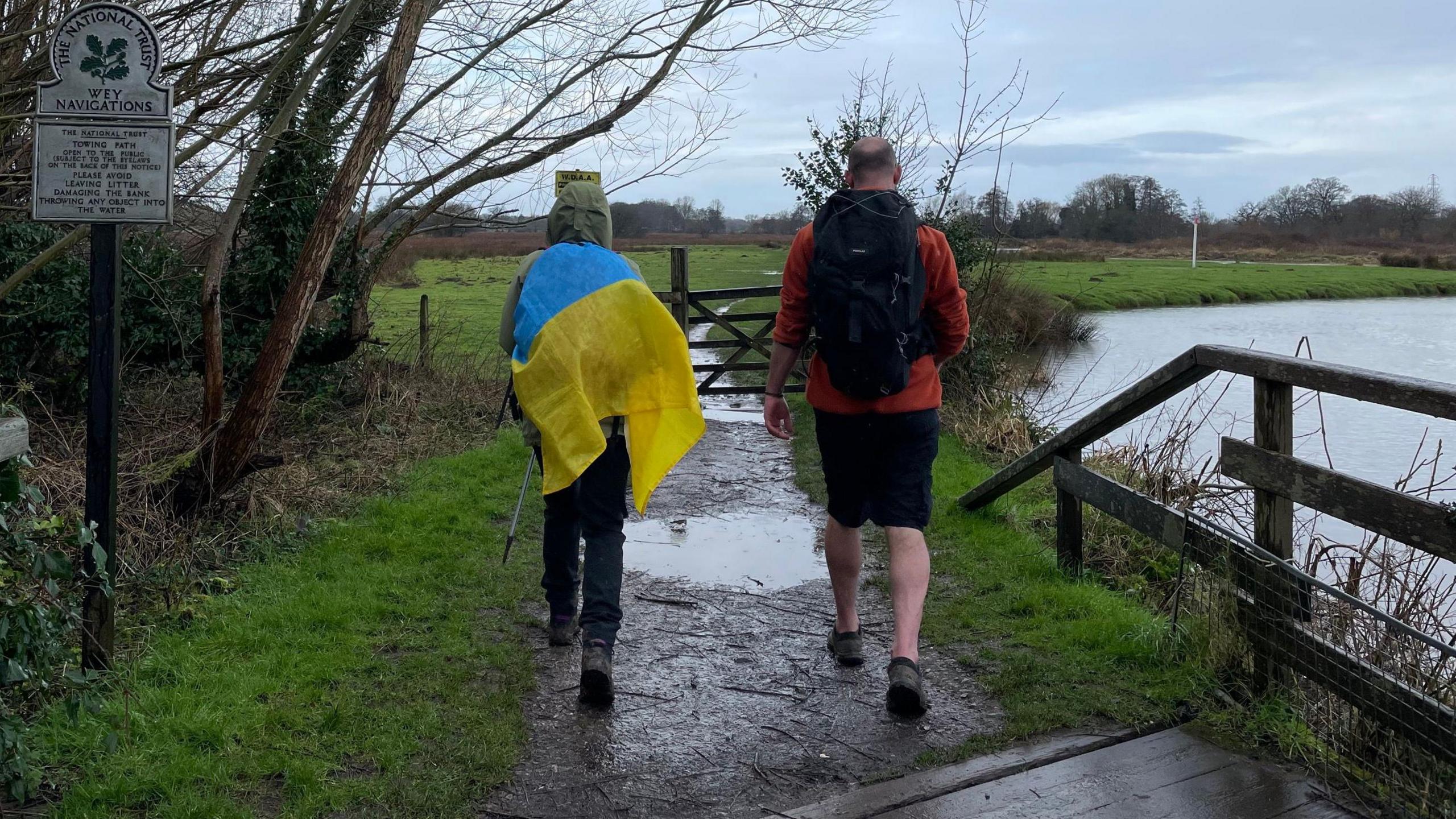 Two people walk along a country footpath with their backs to the camera. One has a Ukraine flag draped on their backpack. There is grass either side of the muddy path. 