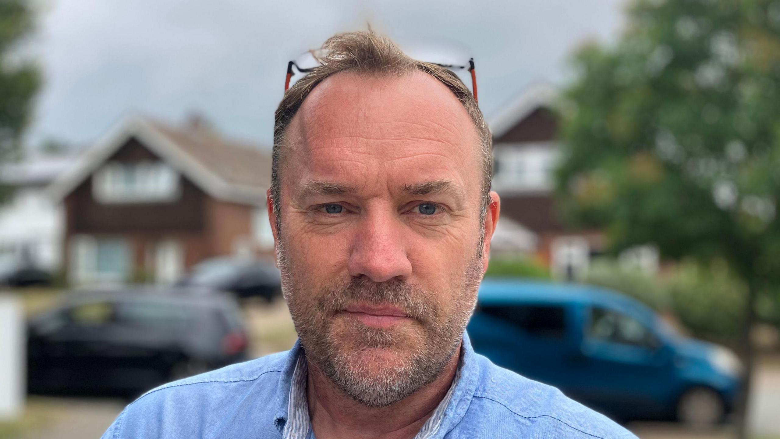 Simon Barrett smiles slightly at the camera. He has thin hair with glasses resting on top of his head. He wears a blue shirt. 