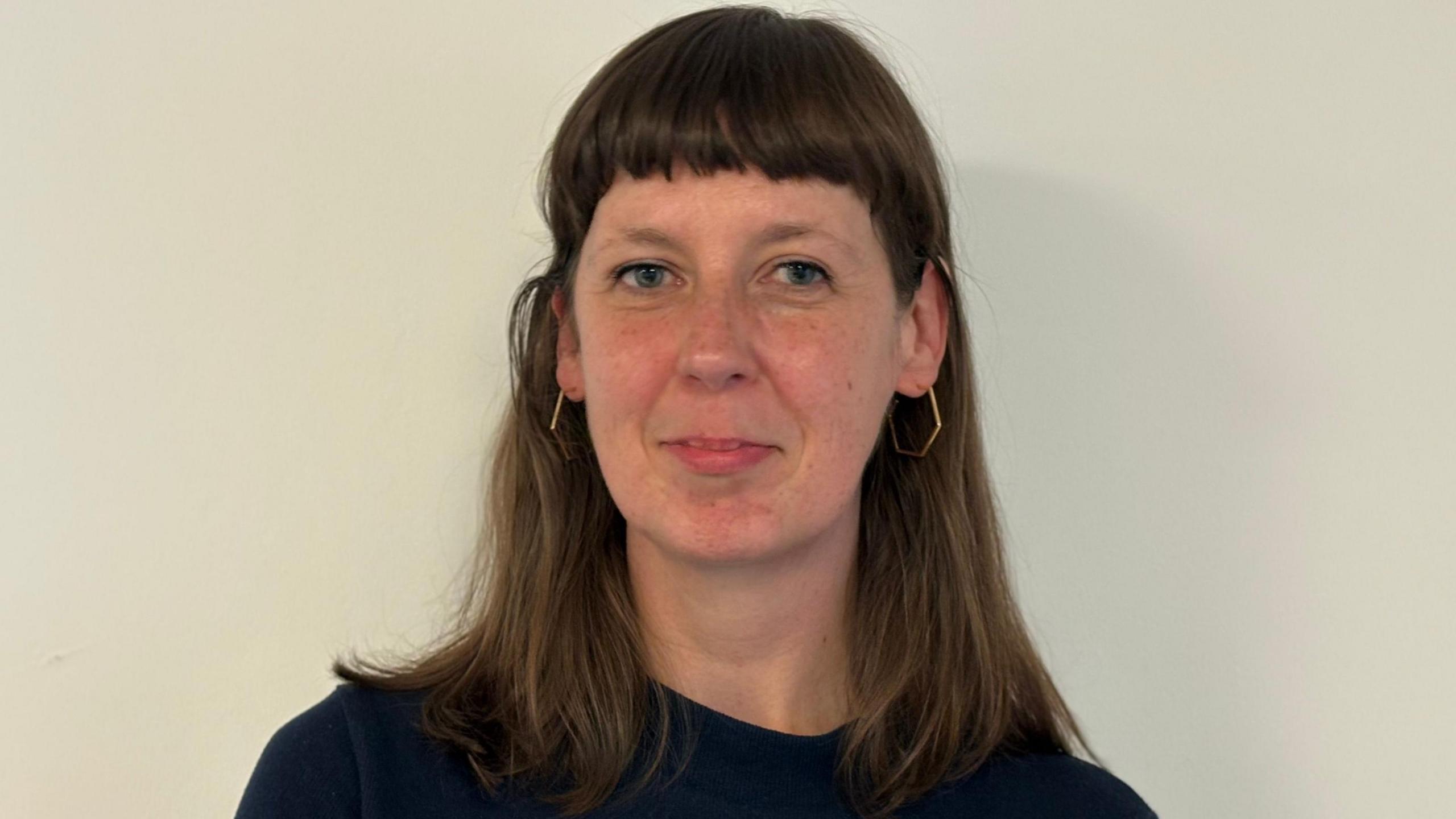 Photo of Sara against a neutral off-white background. She is looking directly at the camera. She has long brown hair and a fringe. Her hair is behind her ears and she wears hexagonal earrings.
