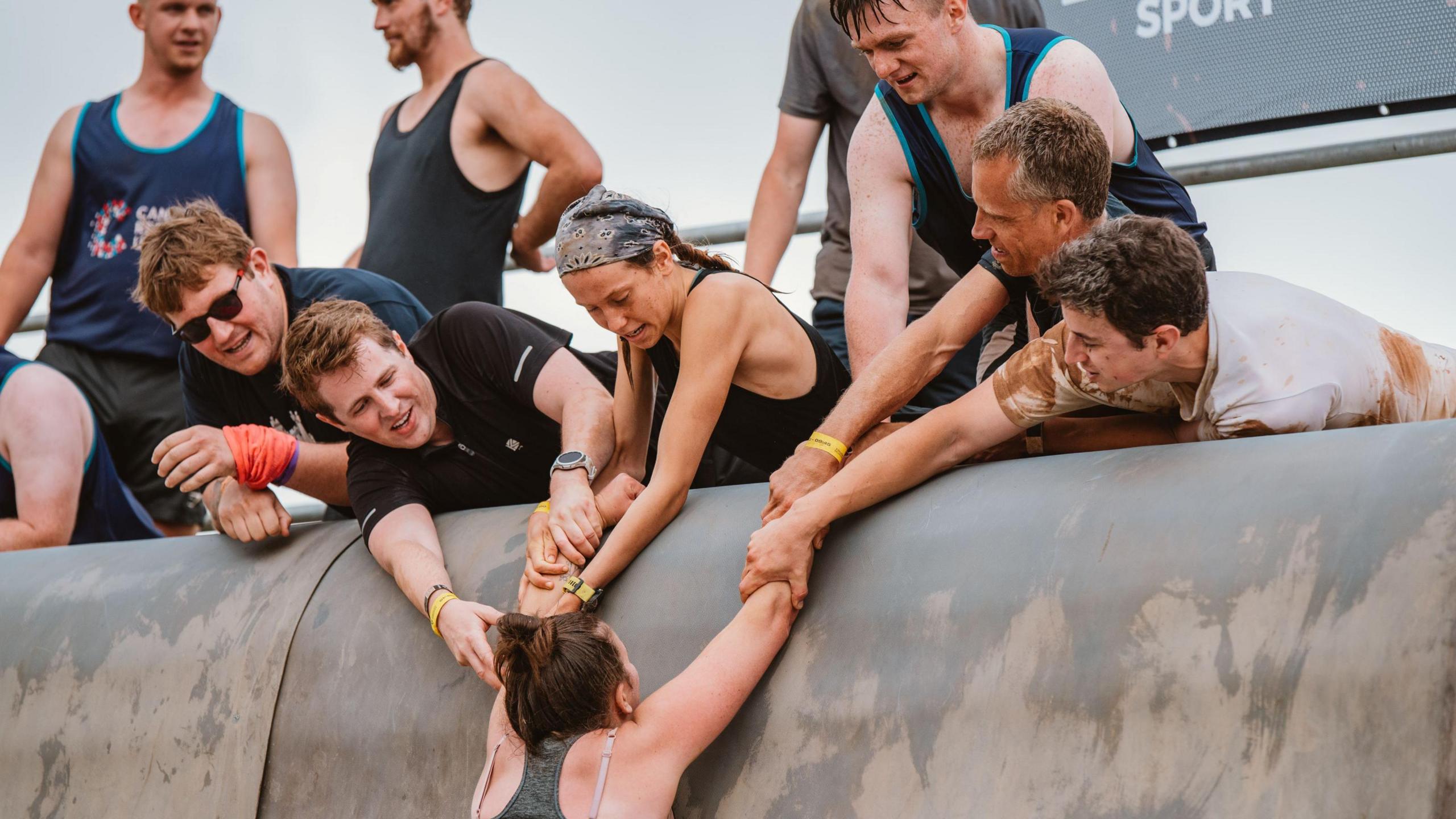 A group of people, some men and some women, pull a female competitor up a steep sided obstacle during a Tough Mudder event on the outskirts of Bristol. They are all wearing dark sports gear and some have mud on them