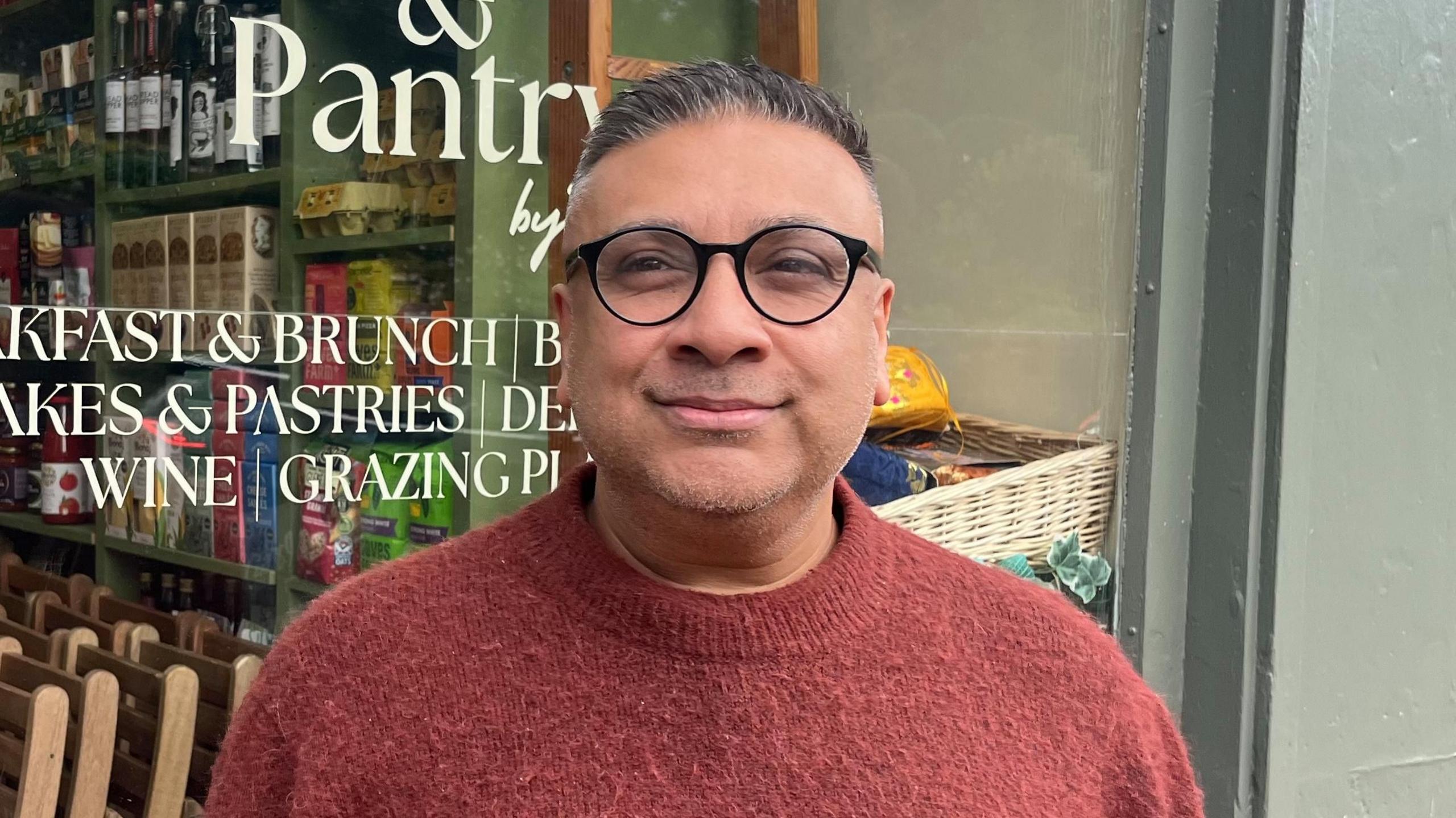 A man with short brown hair, wearing glasses and a red sweater, stands in front of a café and delicatessen. Part of the sign can be seen in the background, as can the boxes and bottles on the shelves.