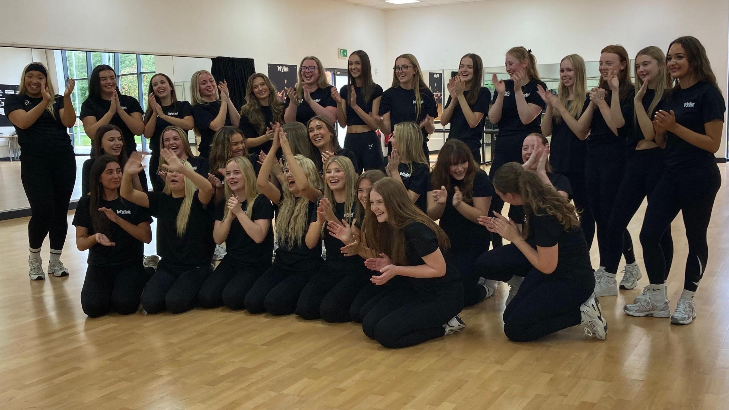 Dancers from Wyke 6th Form College in Hull clap their hands, laugh and smile at the camera. The group is wearing black leggings, a black T-shirt and white trainers. Two front rows are sat on a gym floor and one back row are standing.