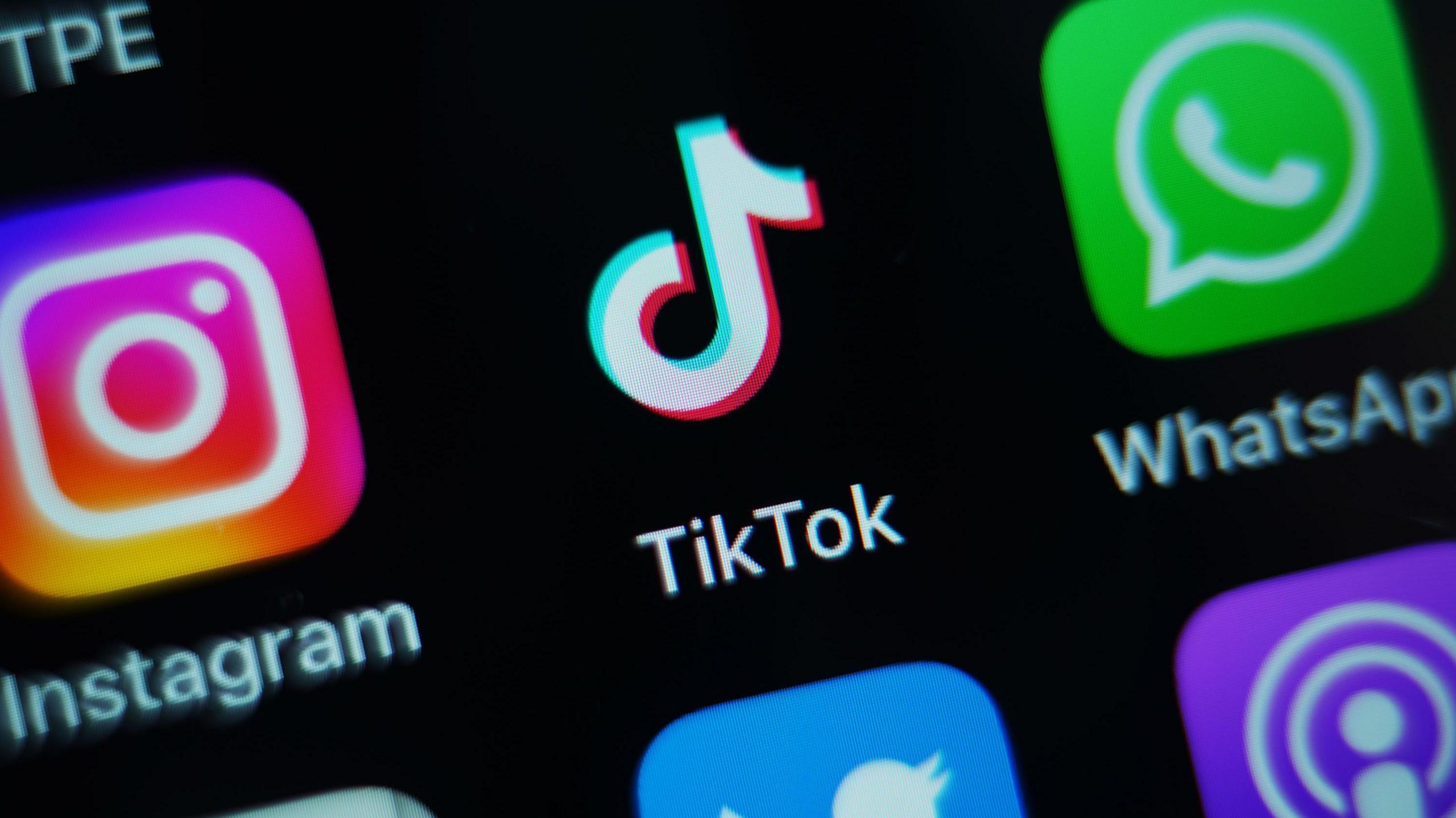 A close up photo of a black smart phone screen showing a number of brightly coloured icons, including the pink and white Instgram icon, the white musical note TikTok icon and the WhatsApp icon which is a green square with a speech bubble and phone handset inside. Each icon has the name of the app below it in white font.