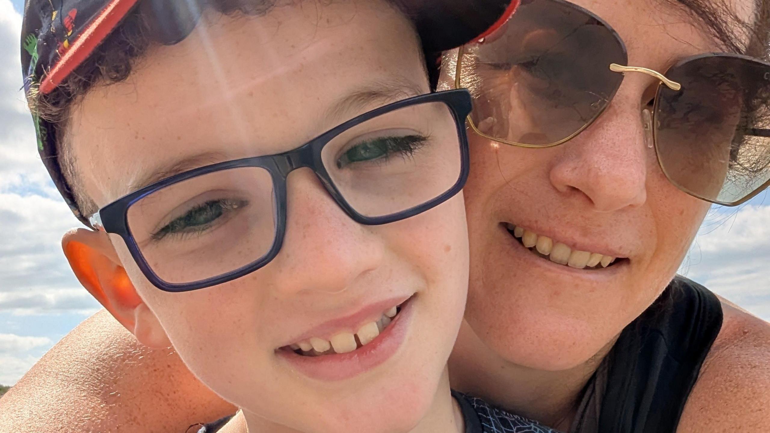 Amy Jones and her son Harry in a candid photo in the sunshine. Amy is behind Harry and has her arms around him. She is wearing a black sleeveless T-shirt and gold-framed sunglasses. Harry is wearing a black T-shirt with line illustrations on. He has glasses with a dark frame. Both are smiling.