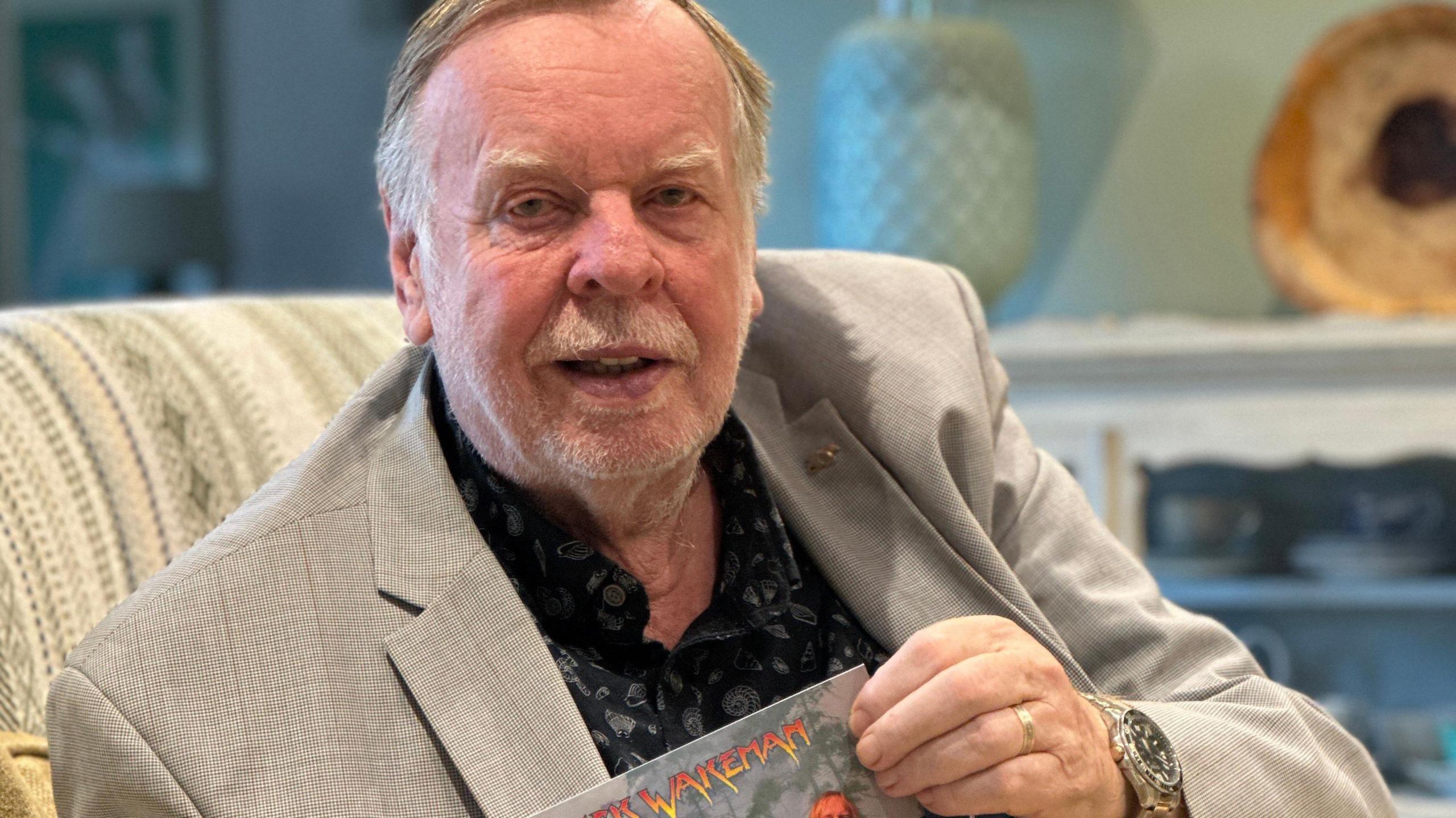 Rick Wakeman, who has short fair hair and beard and is wearing a light coloured suit jacket over a black shirt.