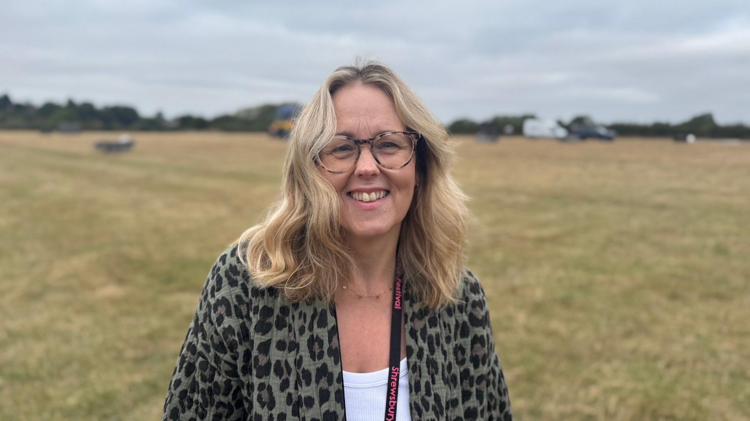 A woman, who has shoulder-length blonde hair, is wearing tortoiseshell glasses and an animal-print cardigan and white T-shirt and has a black and pink lanyard around her neck. She is standing in a field with trees and greenery in the far distance.