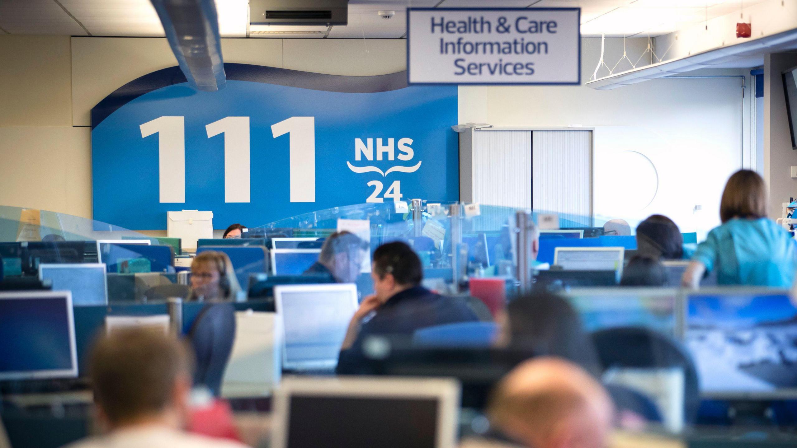 Staff at a desk in a 111 call centre. Many computers can be seen and in the background there is a large blue sign with '111' in large white font.