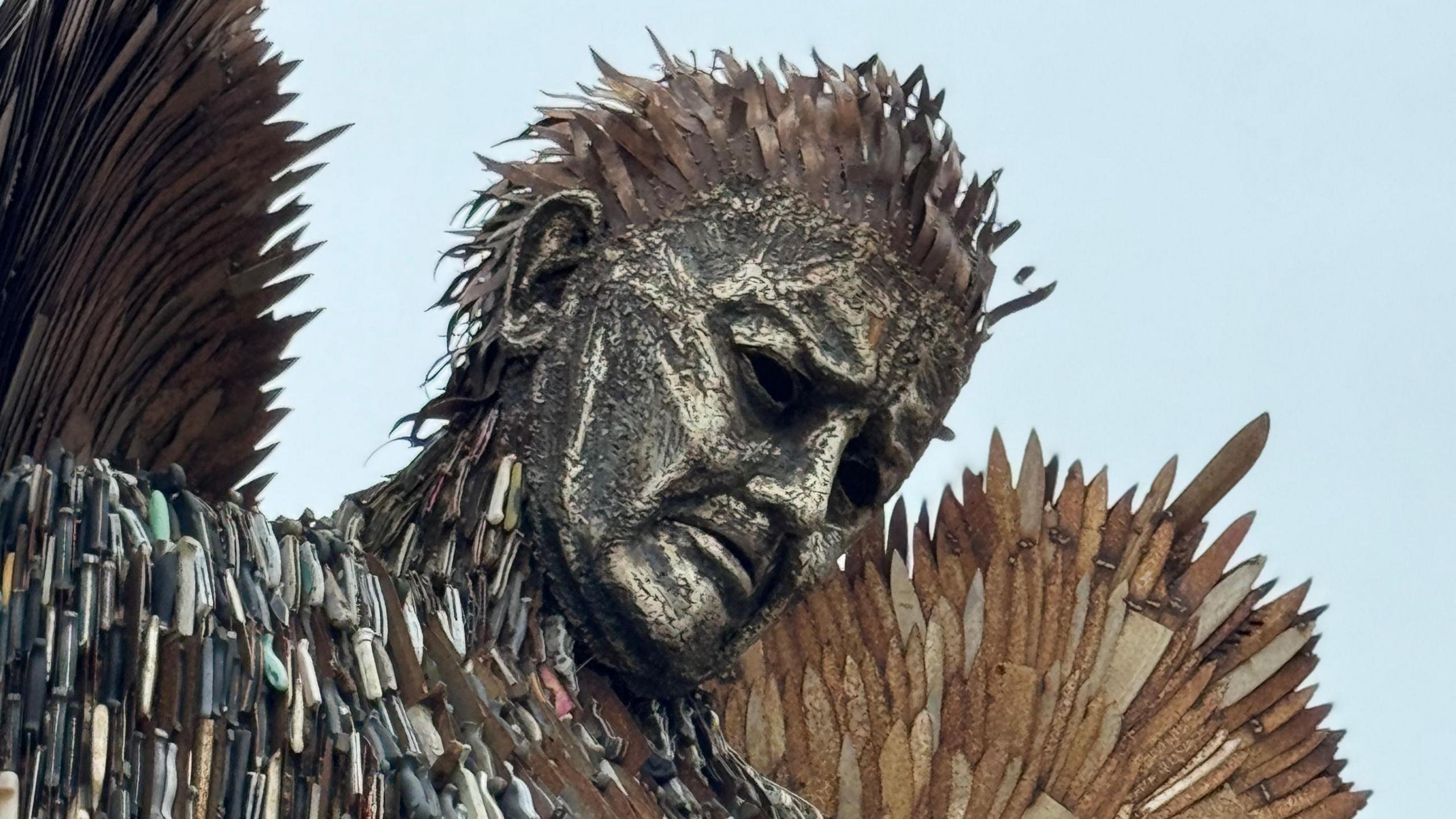 Zoomed in to portrait view of the face of the statue. The face is made of a worn metal, and his eyebrows are lowered to appear upset. Rusty wings made of knifes stretch behind. 
