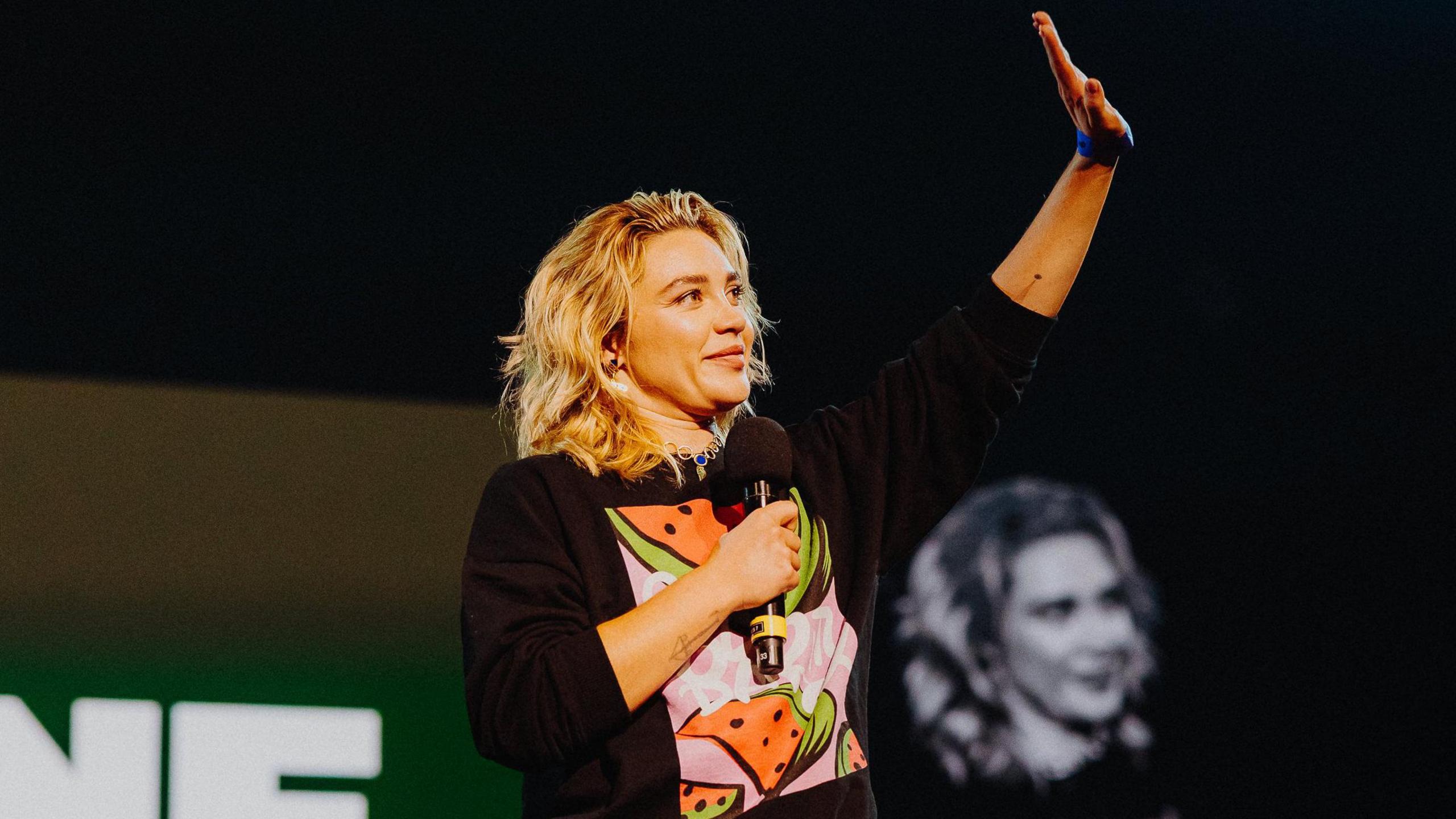 Florence Pugh holds a microphone and waves. She wears a black jumper with a print showing slices of watermelon.