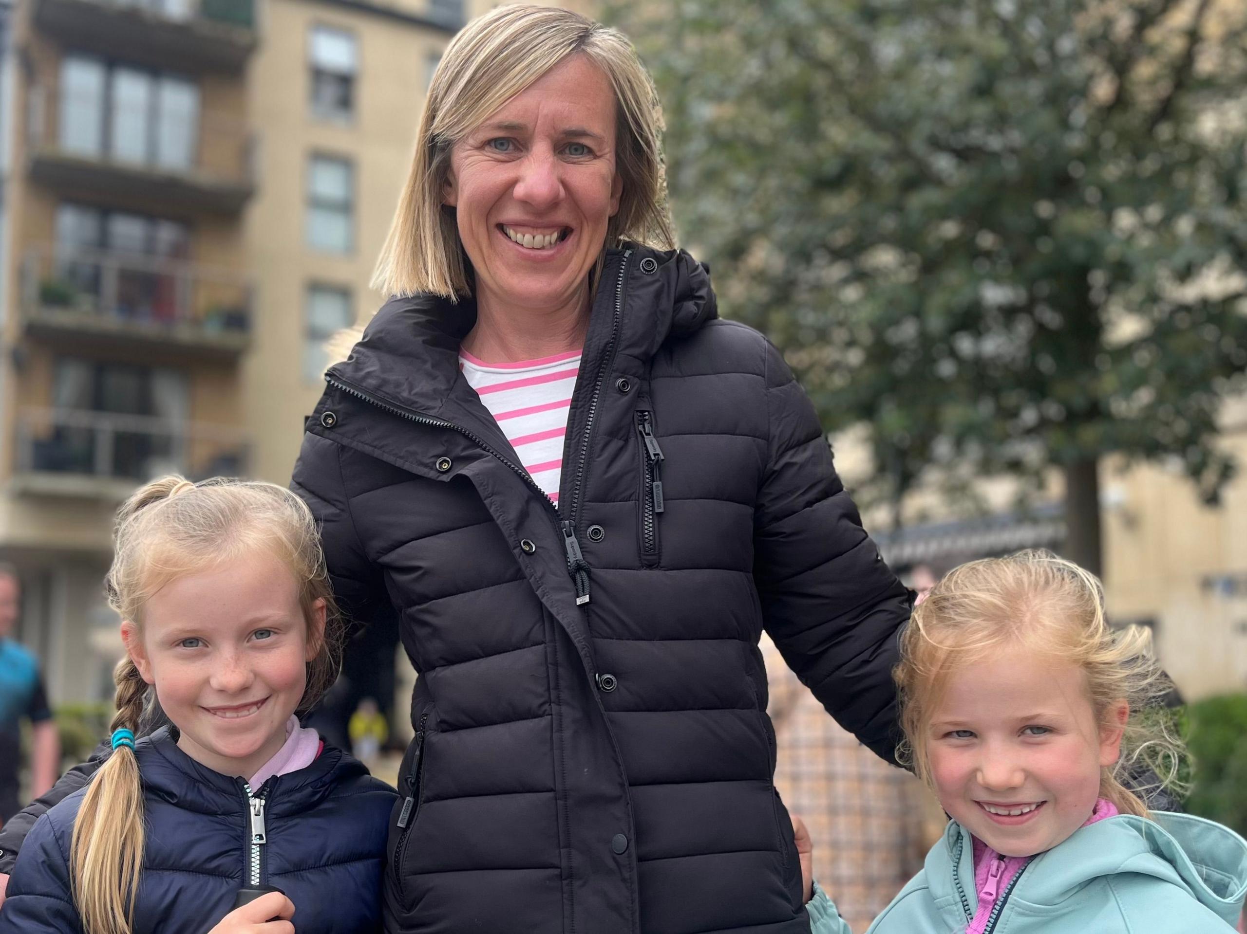 A woman with blonde hair in the centre of the image, smiling at the camera, with her two young daughter either side of her 