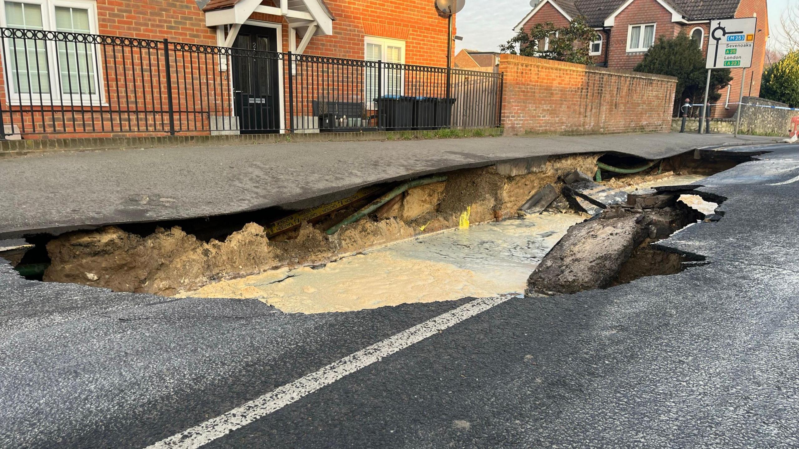 A view of the sinkhole taken from the level of the road surface. It shows the ground beneath the pavement, now exposed by the sinkhole that has opened up in the road. Pooled water is visible inside the hole. A red brick house with black metal fencing sits behind the pavement.