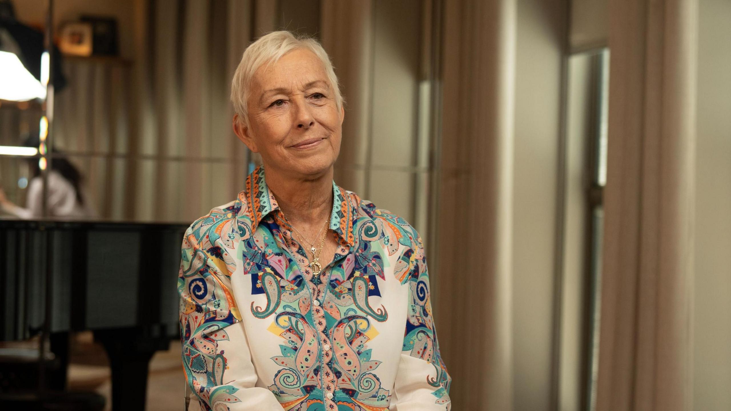 Martina Navratilova, with short white hair, wearing a colourful paisley-print blue and orange collared shirt and a thin gold chain necklace. She's sitting in a room in front of some beige curtains - the background of the photo is out of focus.