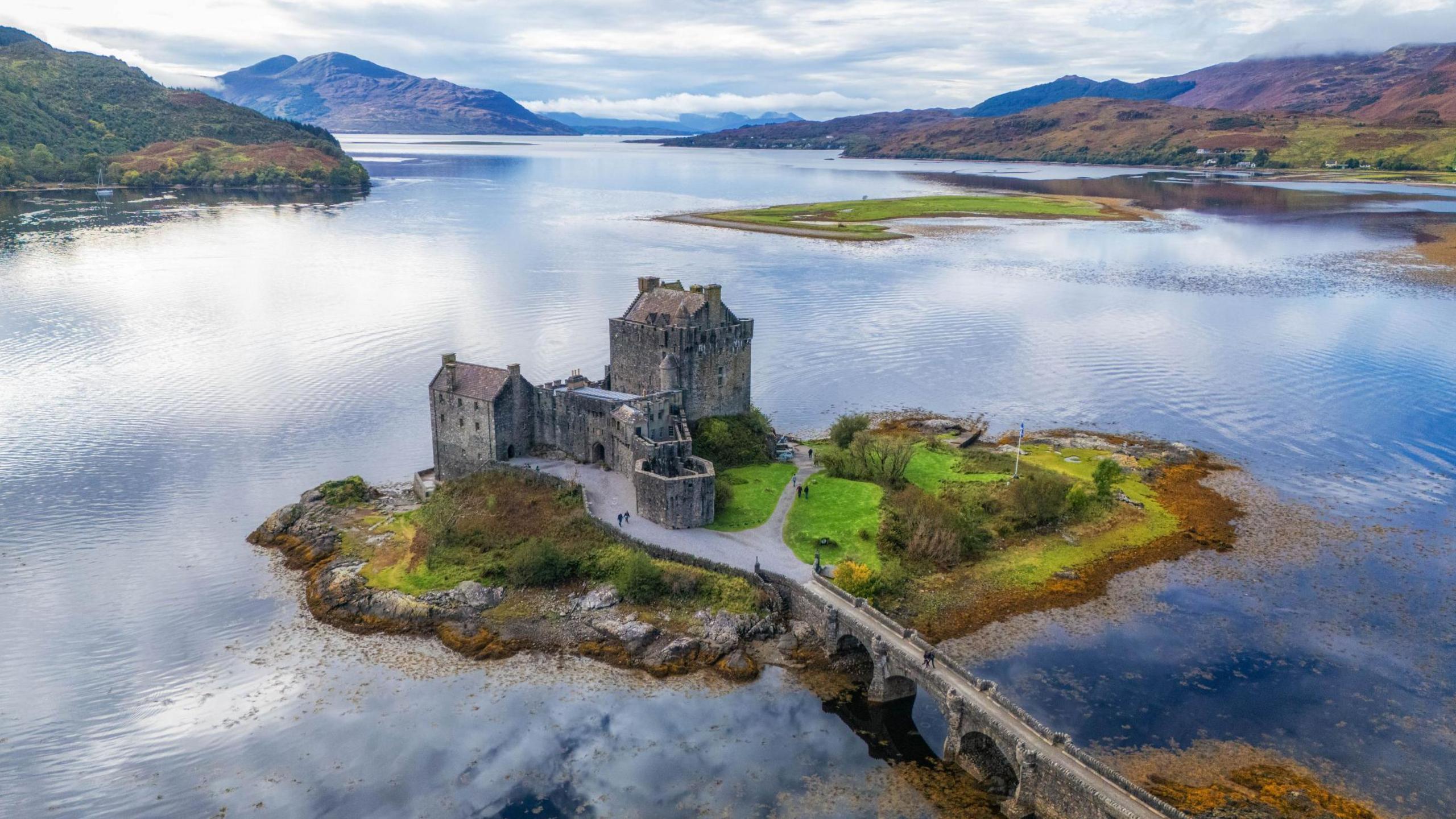 Eilean Donan Castle is a complex of stone-built fortifications on a small island in a sea loch. It is connected to the shore by an arched, stone bridge. The loch is flat calm and in the distance as hills and mountains.
