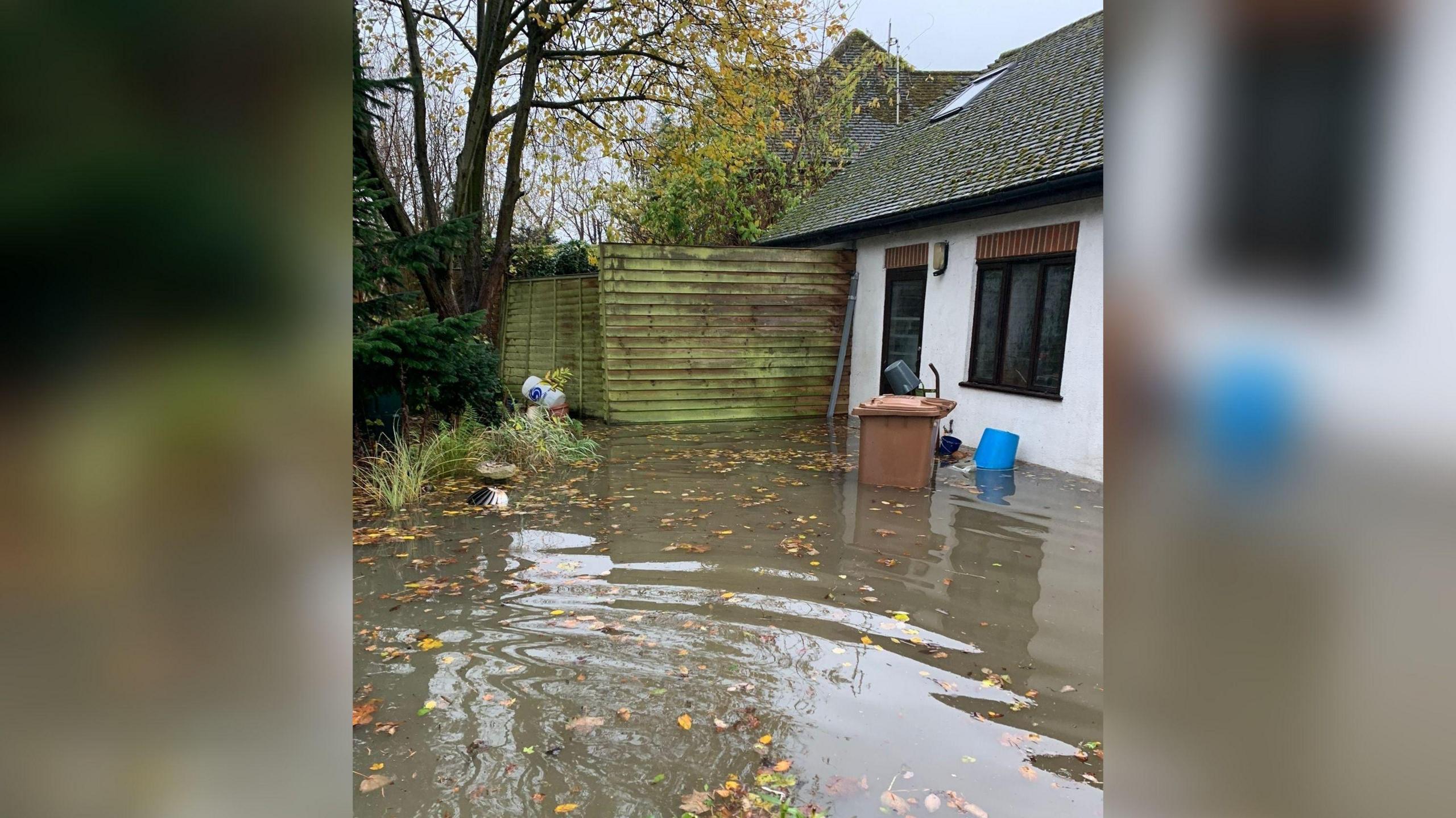 The outside of a house where there is single storey building with water reaching up the walls, and a brown wheelie bin outside which has water reaching about halfway up it.