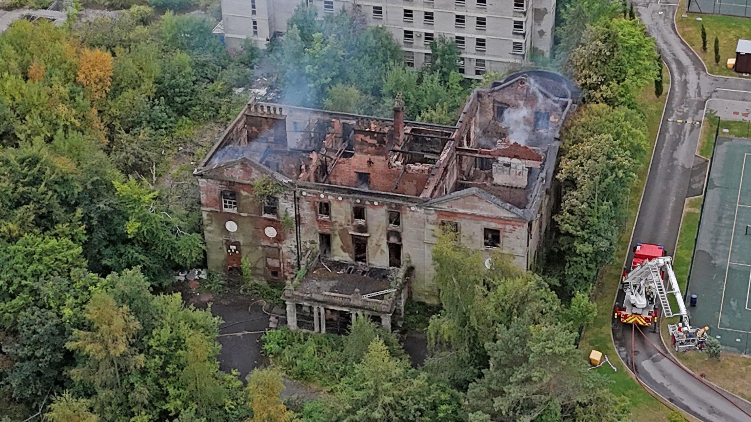 An aerial shot of Woolton Manor. It is an old house surrounded by trees. The house is still smoking and the roof has been completely destroyed.