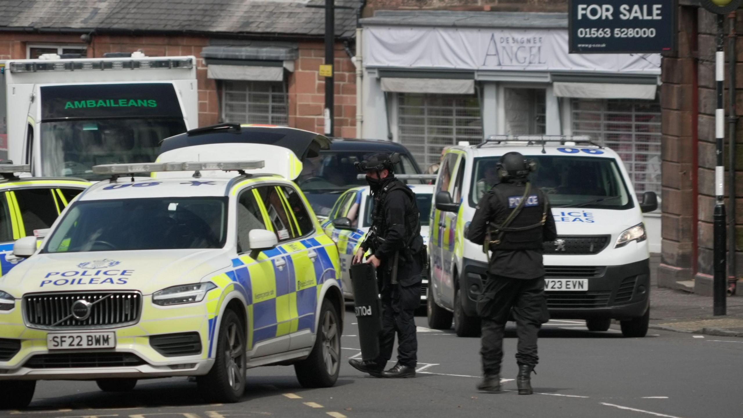 Two armed police officers in bullet-proof vests and wearing black helmets stand next to three police cars and a police van on the street