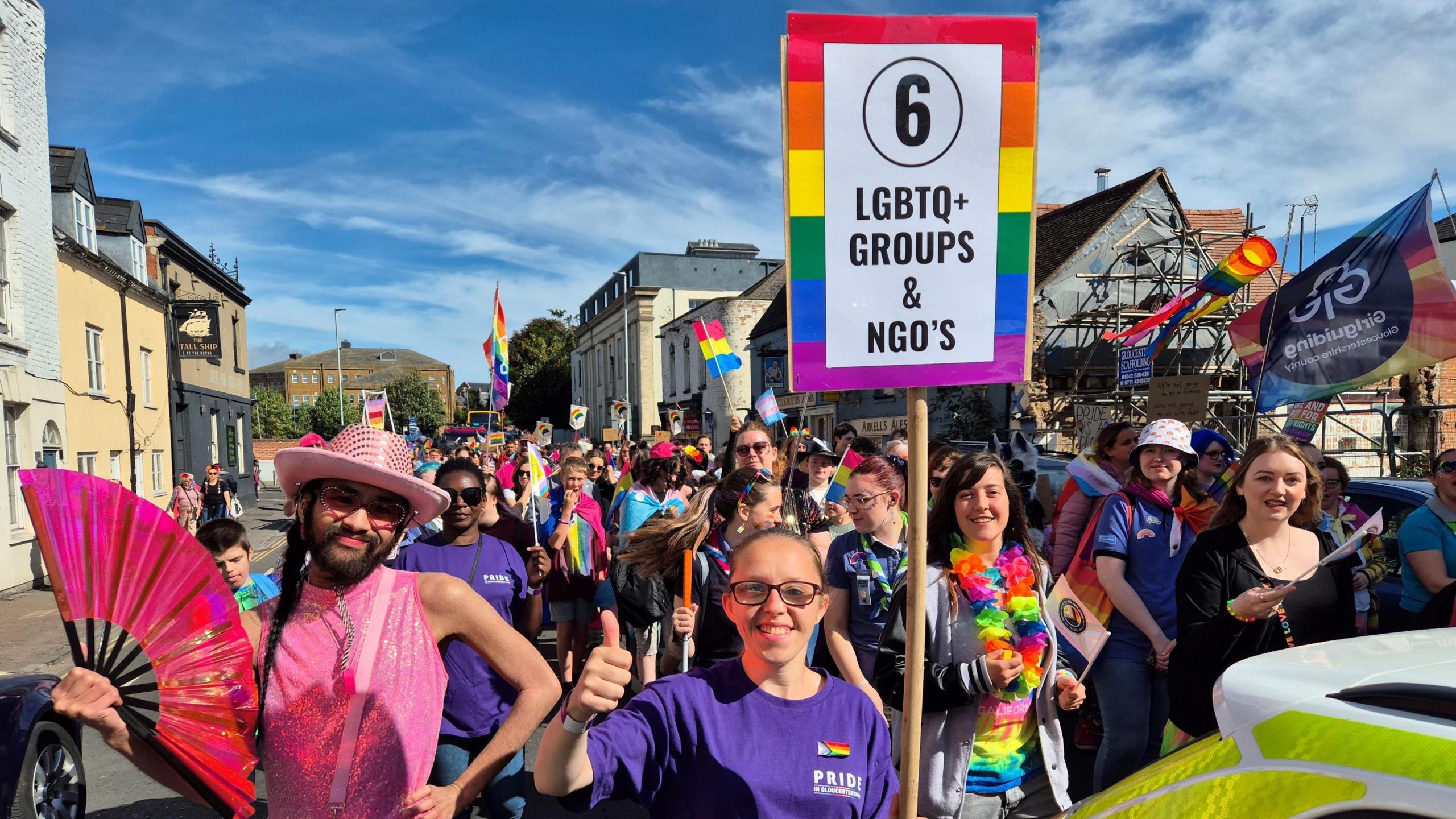 People marching down the straight for Pride in Gloucestershire 2024, holding rainbow flags and other items with a board at the front saying LGBTQ+ Groups & NGOs