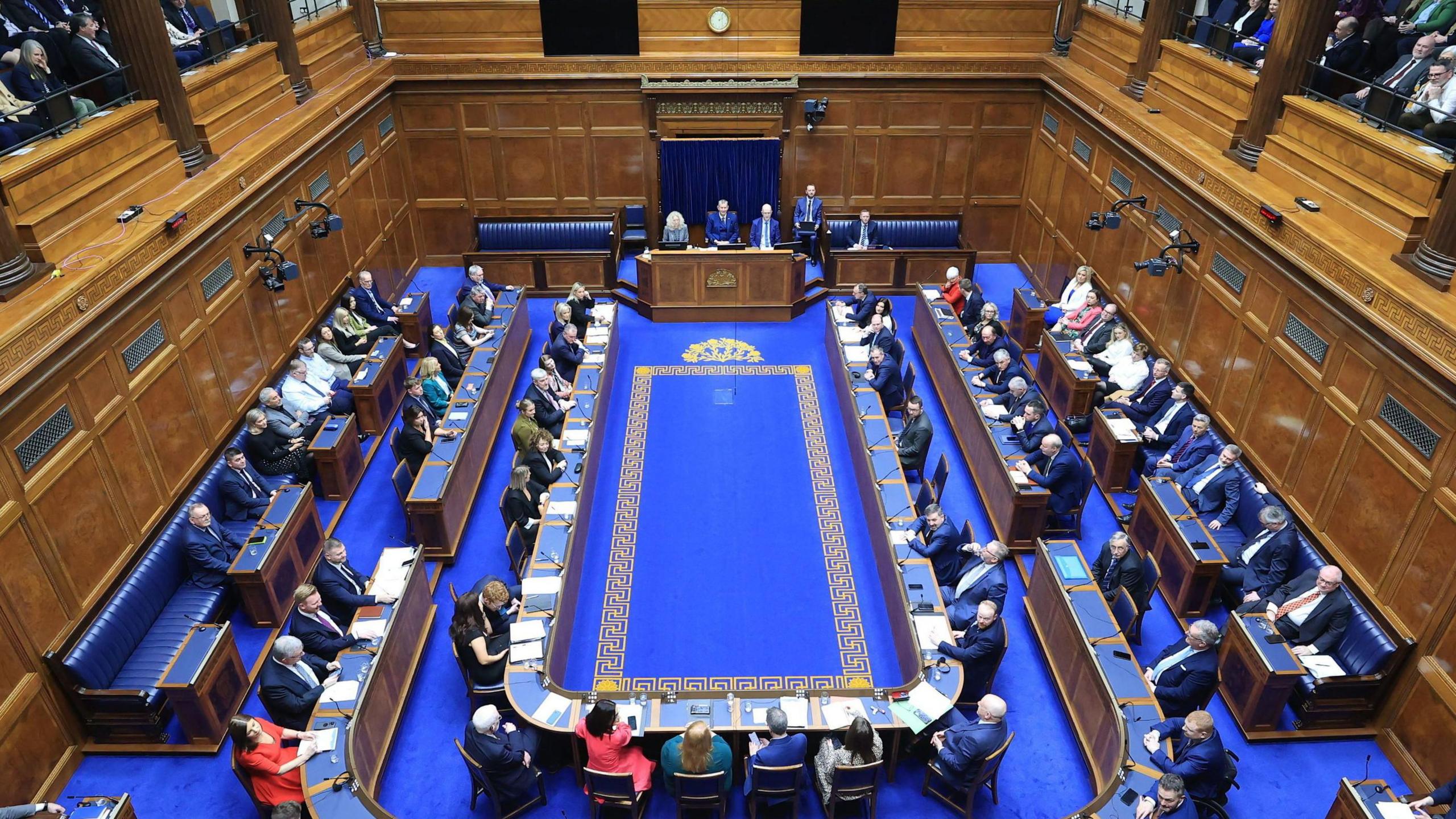 General view of the chamber of the Northern Ireland Assembly in Parliament Buildings, Stormont. The walls are panelled and the carpet is blue.