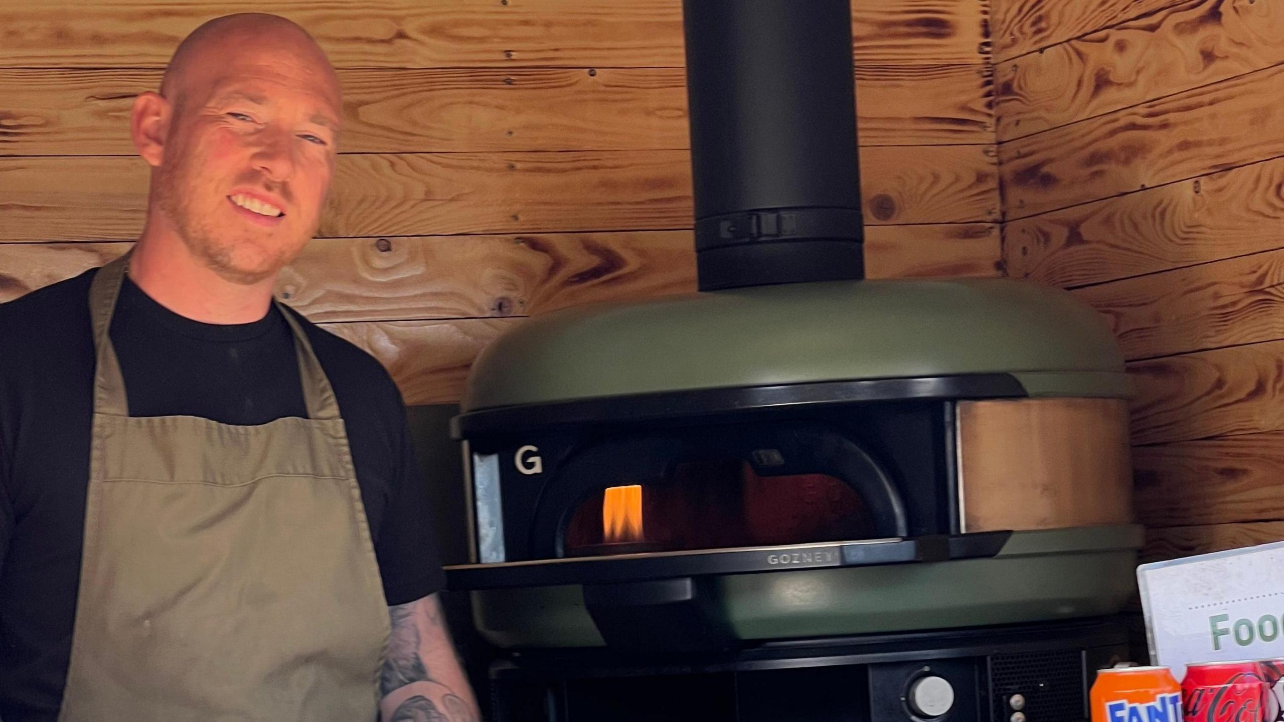 Dan Puddle in his mobile pizza van with a small pizza-oven in the background. He wears an apron and is smiling.