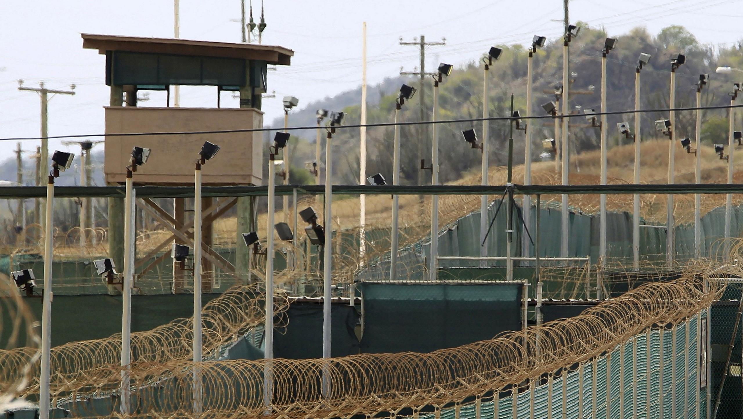 Aerial photo the prison at Guantanamo Bay shows high fences topped with barbed wire, several cameras on poles and a watch tower. There are trees and brush in the background as well as another fence