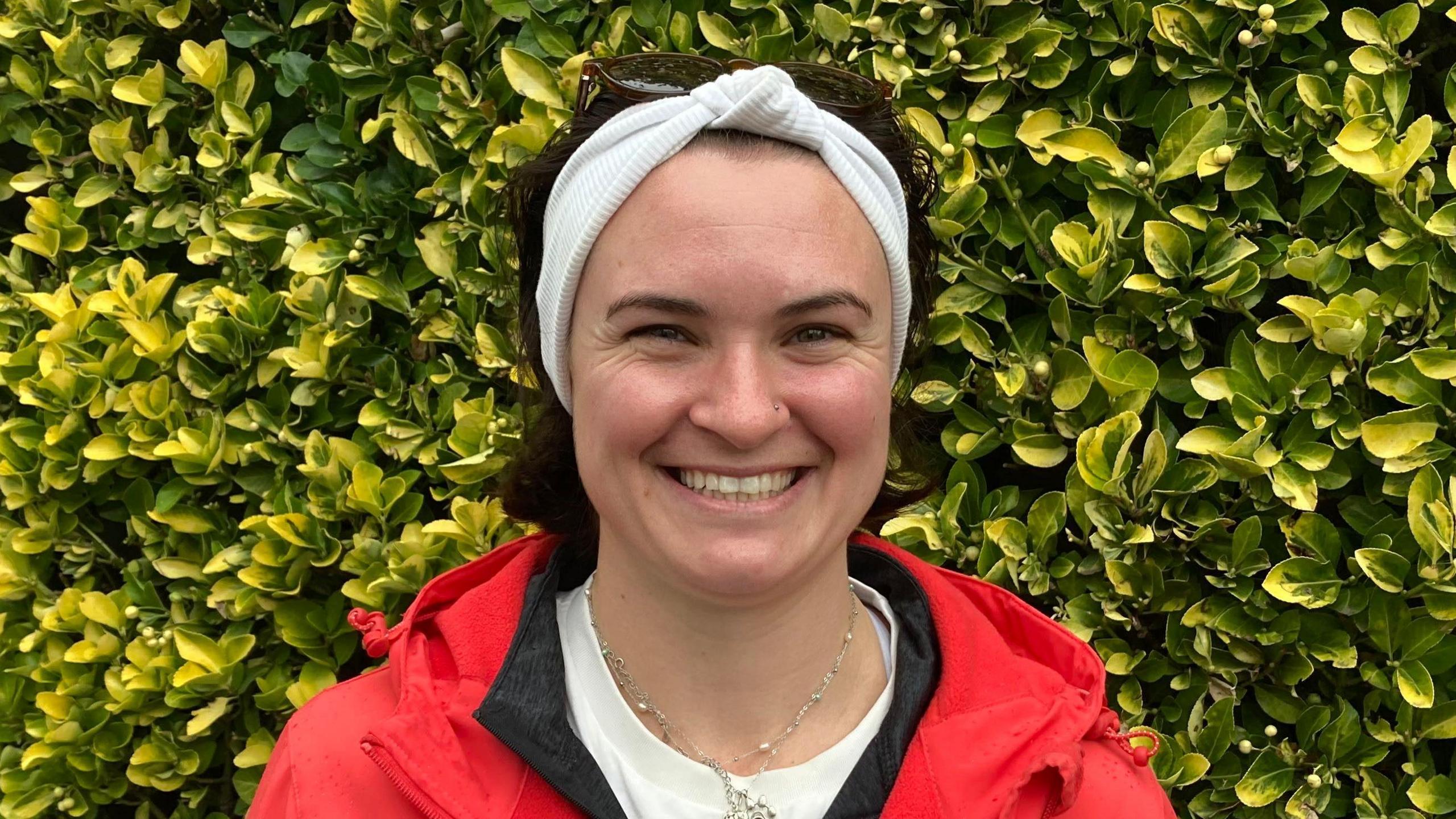 A picture of a lady smiling while wearing a white headband and red coat. She is pictured in front of a hedge.