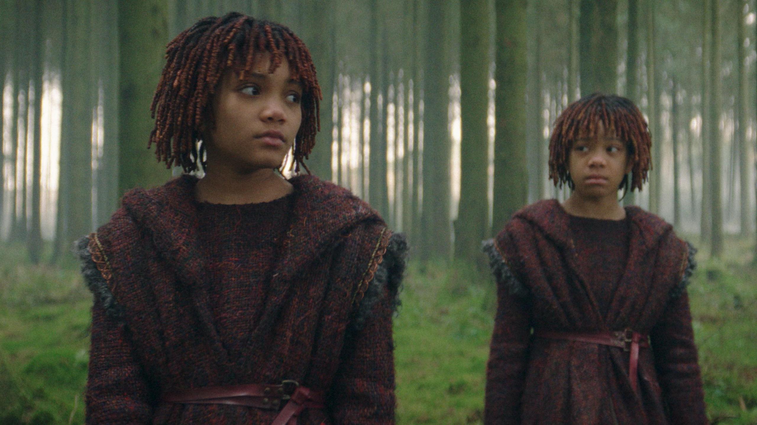 Two young children in robes stand in front of a wooded area.