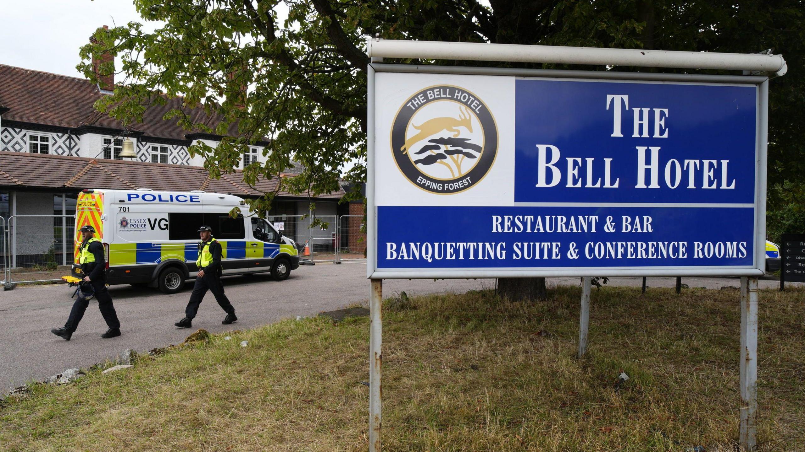 Two police officers walking next to the hotel, which is surrounded by a thin metal fence. A police van has also parked next to the building. In the foreground is a large blue sign which reads 'The Bell Hotel'. 