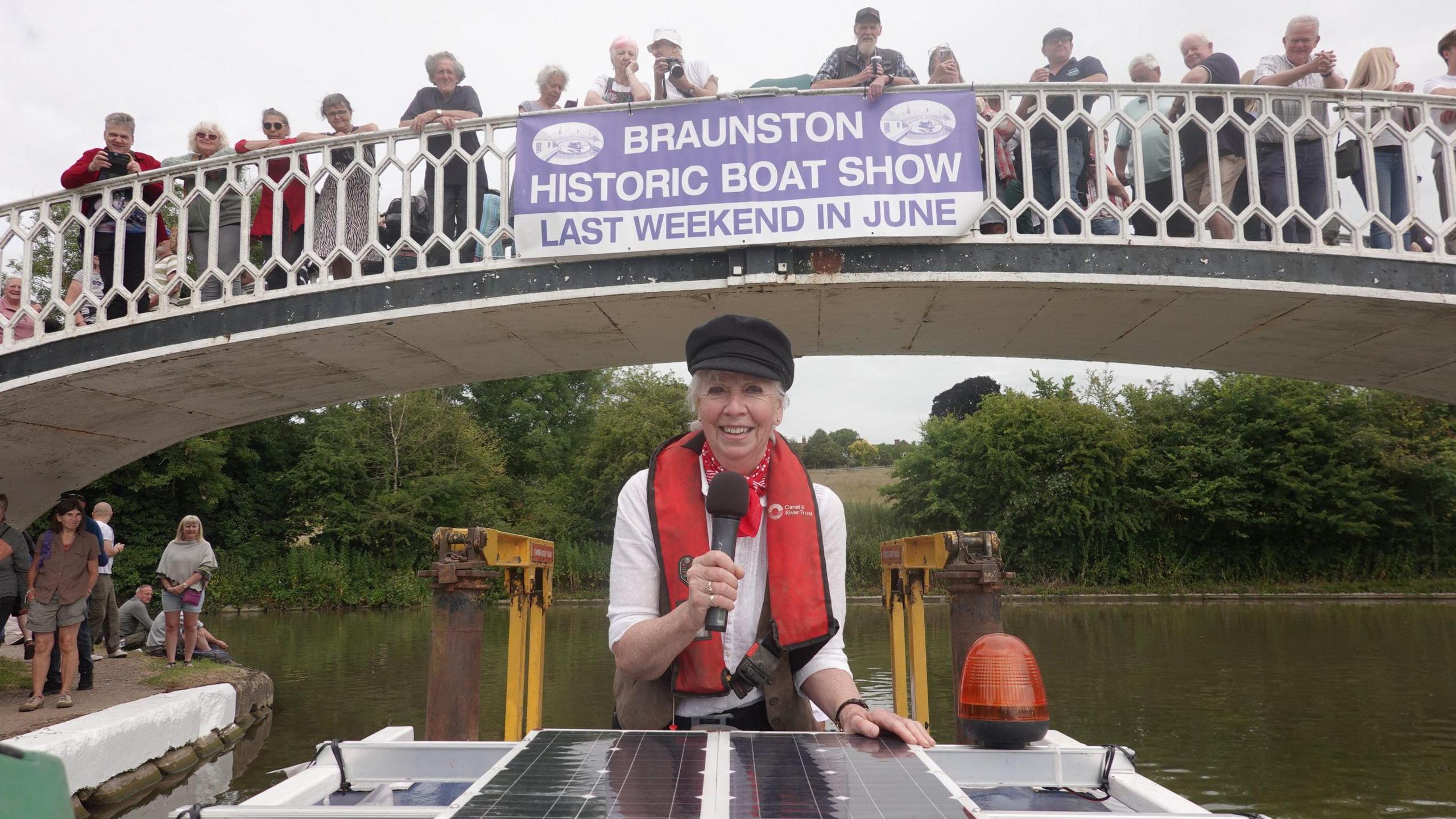Lady Sheila is wearing a black cap, a red life jacket and a white top. She is holding a microphone while riding at the back of a boat which has just gone under a bridge with white railings. There are several people on the bridge, on which a sign saying BRAUNSTON HISTORIC BOAT SHOW is hanging.