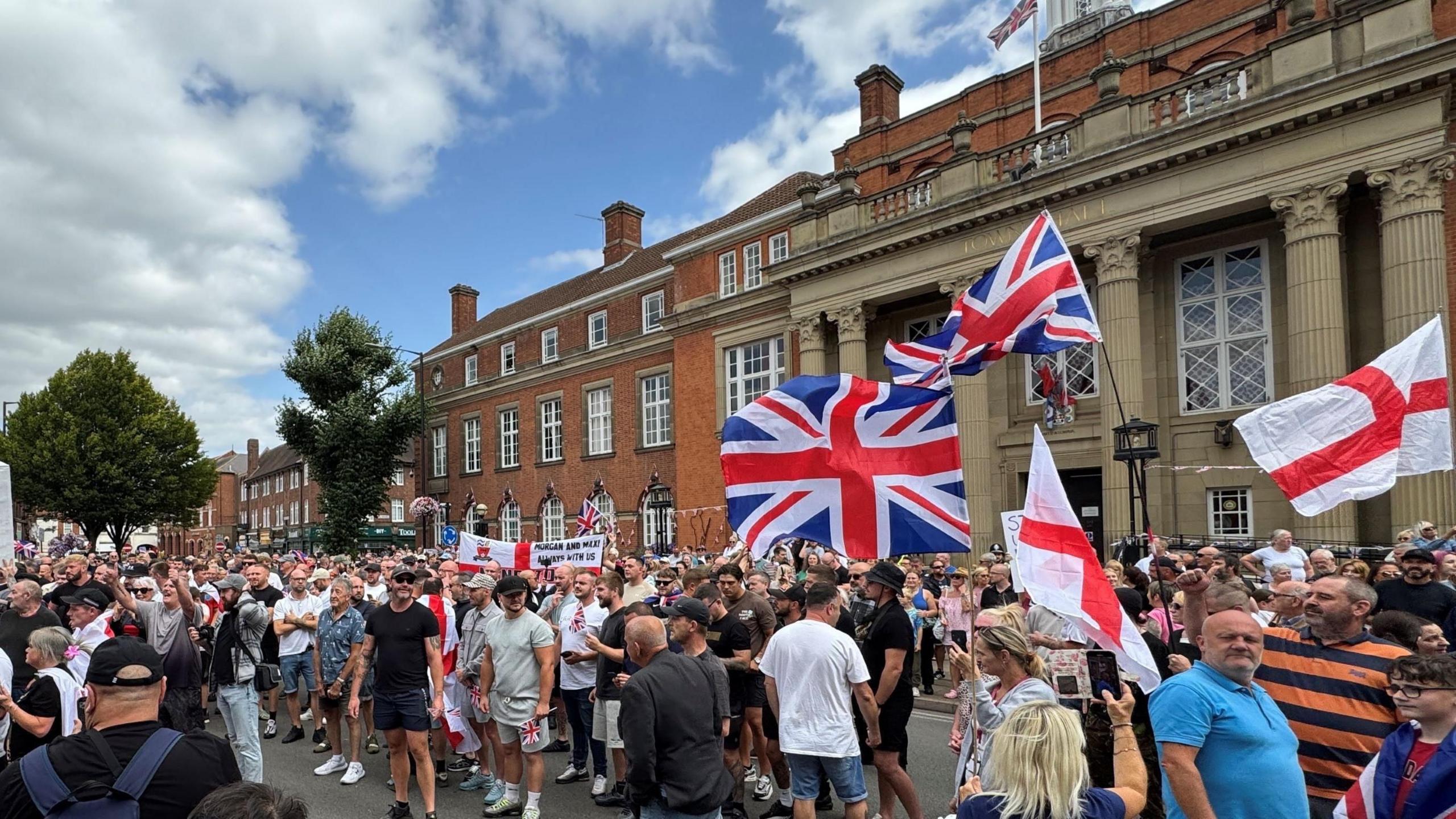 A large group of people with some holding the flag of England and other waving the union jack. They are on paving by large, red brick and stone buildings.