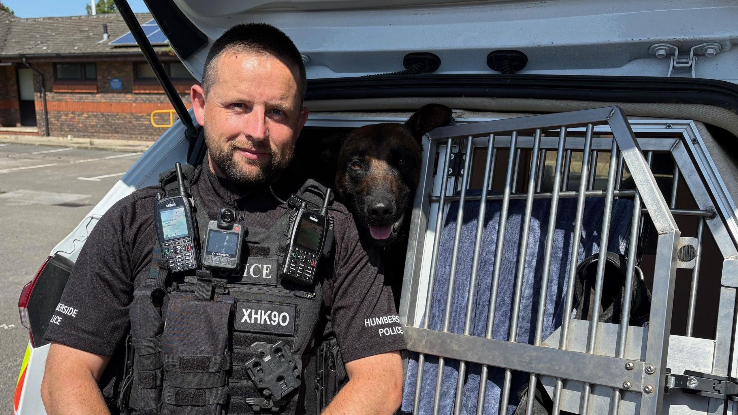 A police officer next to a cage, in the back of a police car, containing a brown and black Belgian Malinois