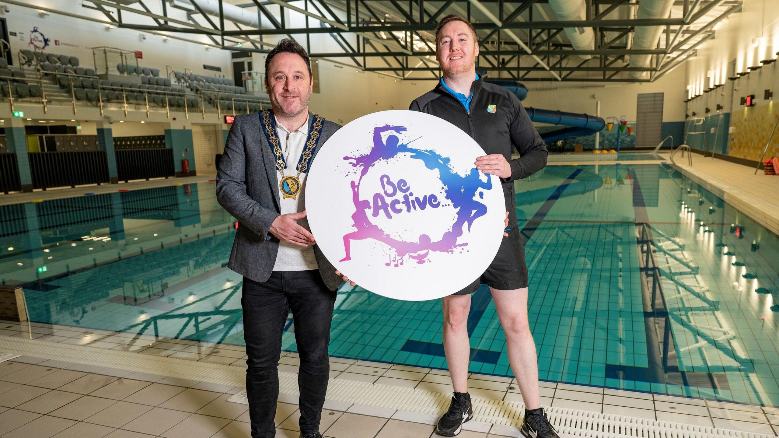 Councillor Pete Byrne and Newry Leisure Centre supervisor Sean McElvanna stand at the side of Newry pool holding a large, round sign that says "Be Active." Pete has short black hair and is wearing his chains of office, a grey jacket and black trousers.  Sean  has short, reddish hair and is wearing a black sports top and black shorts. 