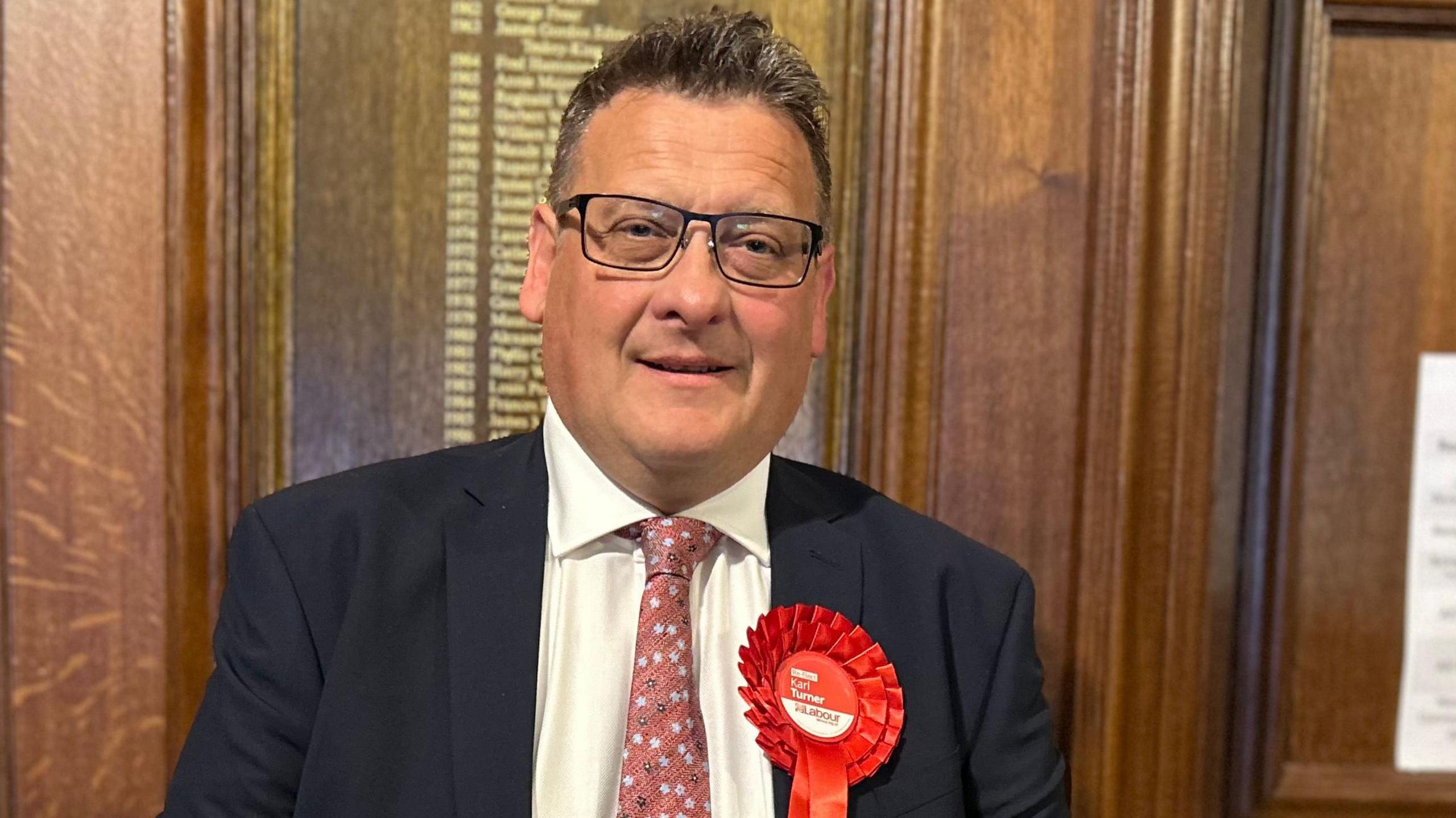 Karl Turner the succesful Labour candidate for Hull East pictured on election night in 2024 wearing a dark suit jacket, white shirt, and patterned pink tie, standing indoors against a wooden paneled wall. A large red rosette with the word ‘Labour’ and smaller text is pinned to the left side of the jacket. 