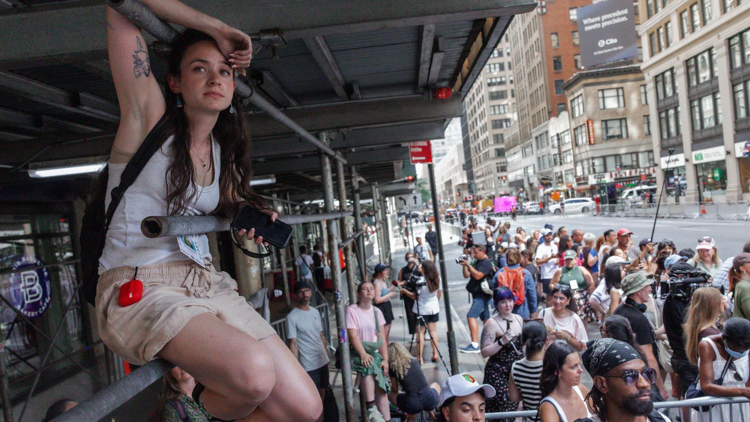 A woman sits on scaffolding, while a crowd of onlookers gather below