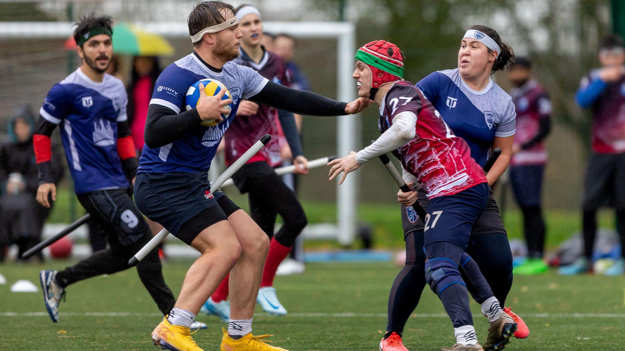 A player cradles a ball while trying to guard it from an opponent. Their hand is outstretched and nearly touches the other player's shoulder. Three other players can be seen in the background coming to their aid.