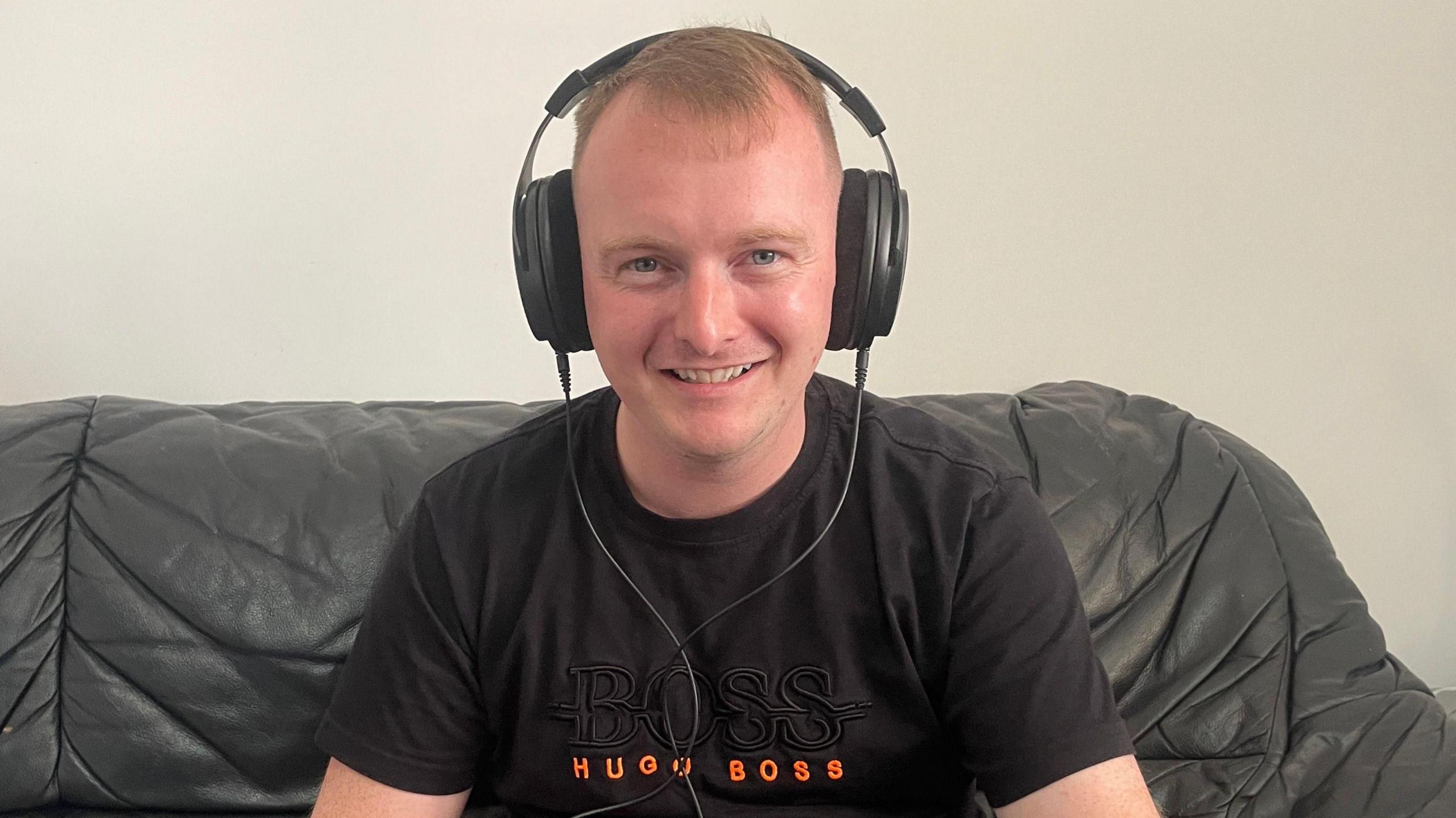 A man wearing large black headphones and a black t-shirt, while sitting on a black leather sofa.