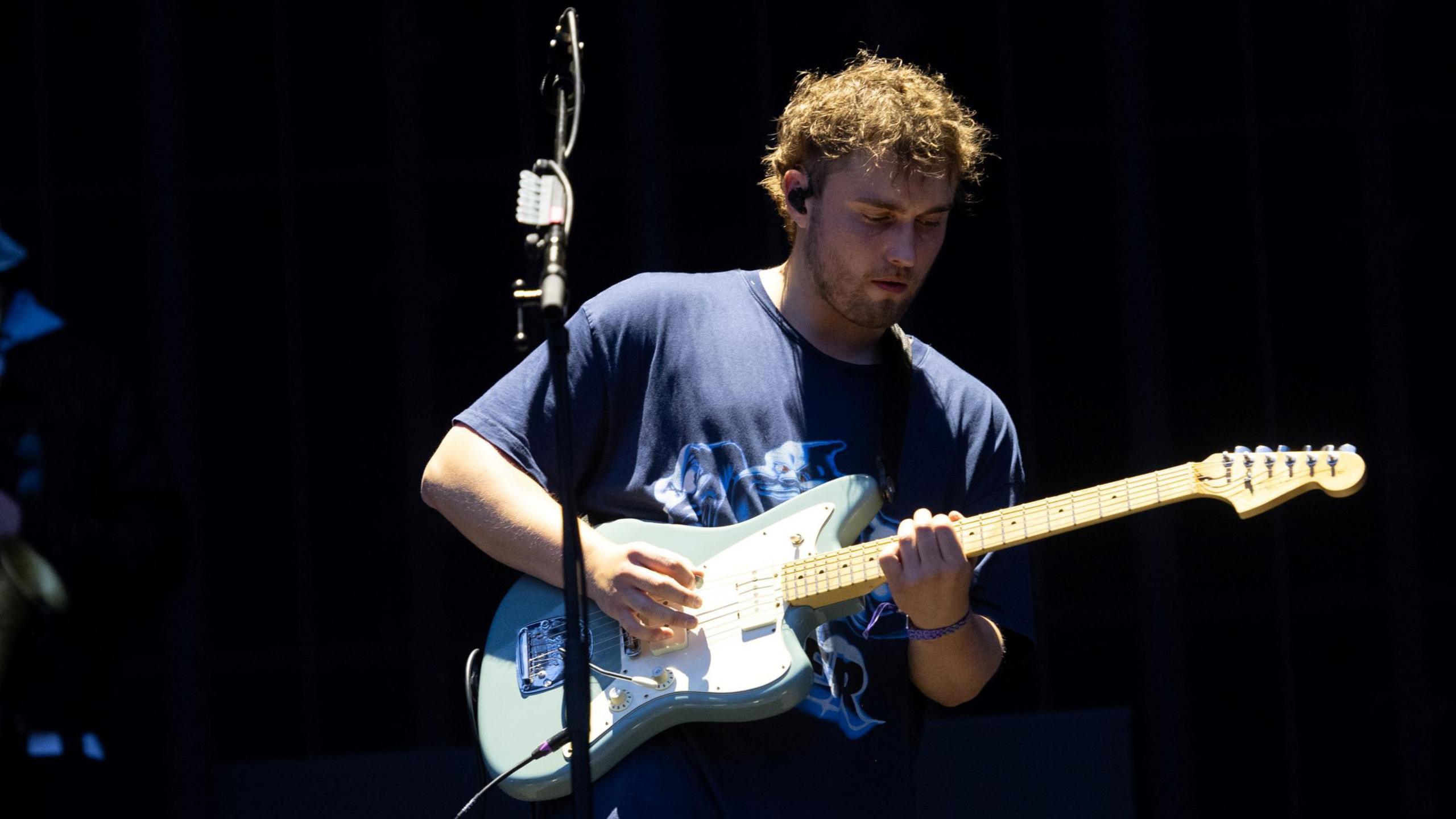 Sam Fender on stage, playing a blue and white electric guitar. He wears a blue T-shirt and the staging behind him is dark.