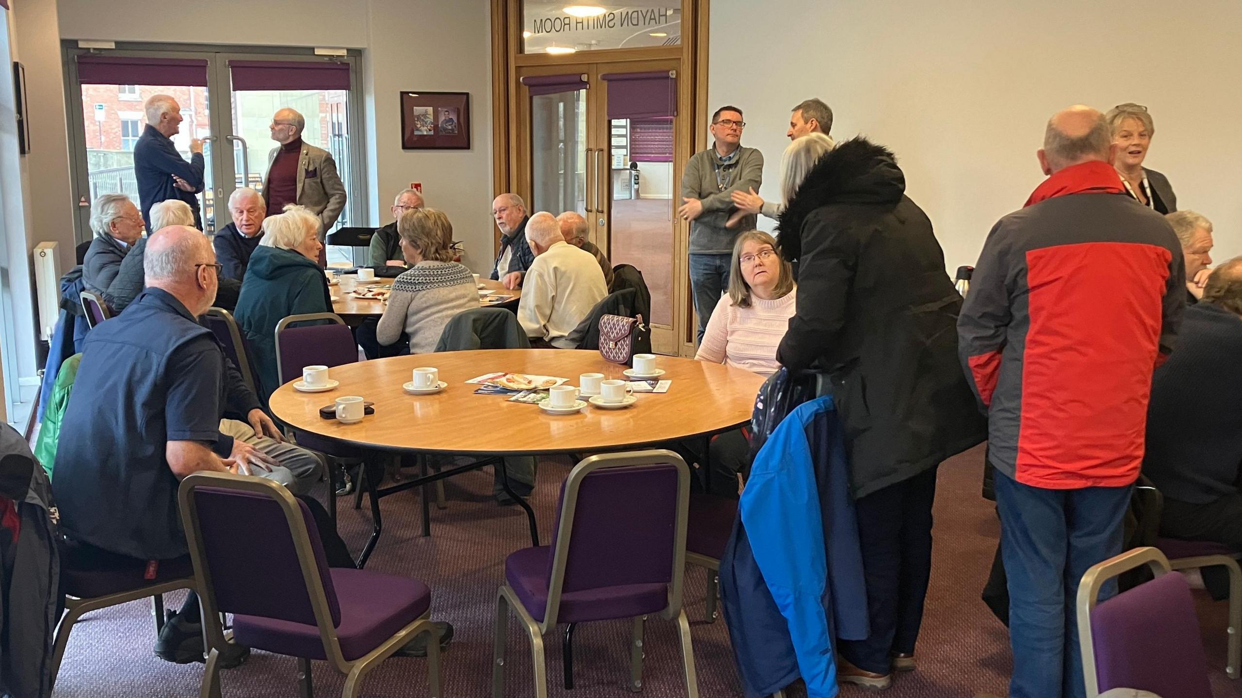 Men and women sat round circular tables and purple-backed chairs inside a cream-coloured venue room, enjoying teas and coffees. Others are standing up wearing coats.