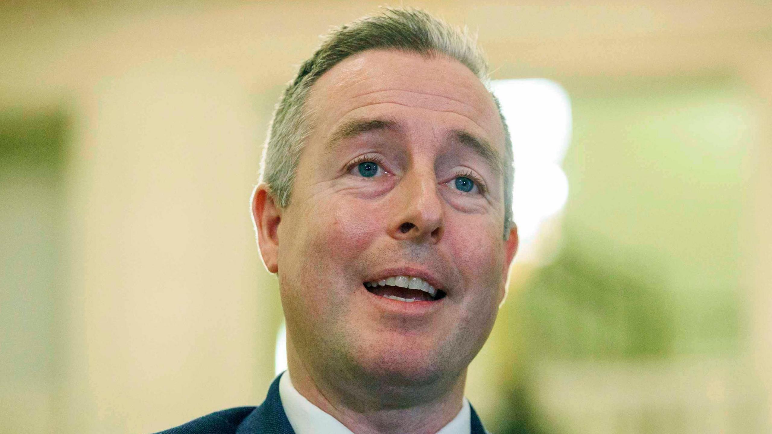 Paul Givan is standing inside the Stormont Building, he is speaking to press. He has short white hair and is wearing a dark suit.