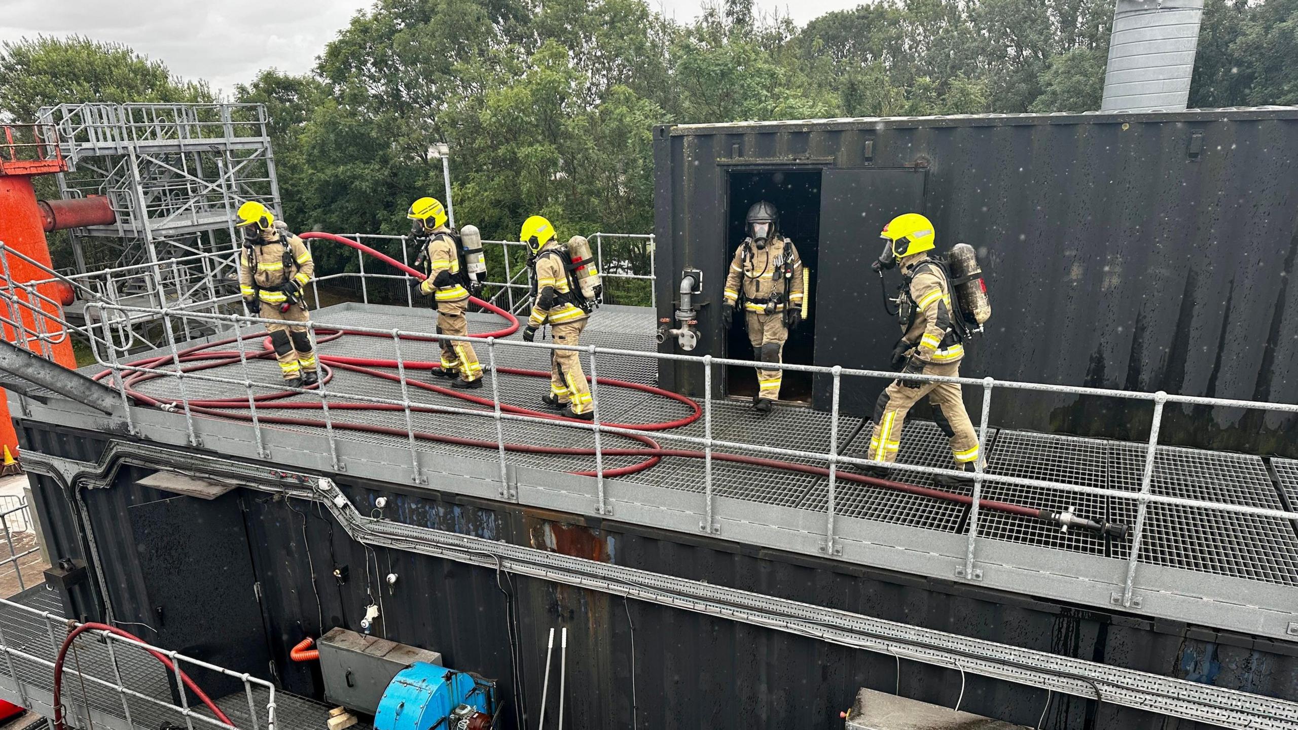 Five firefighters are walking on top of a shipping container with a red hose.