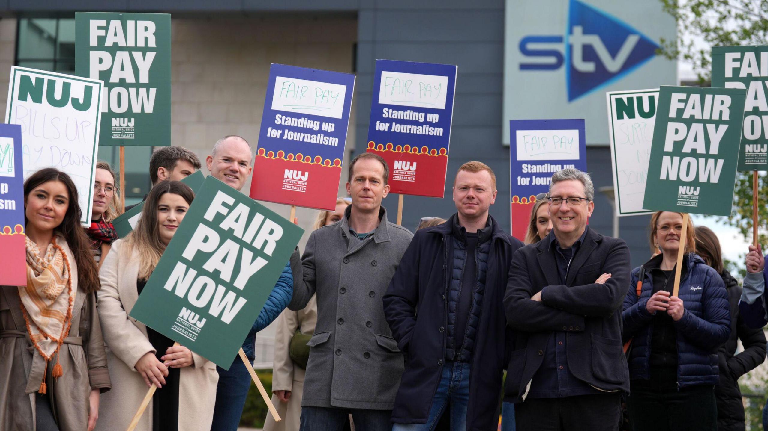 Several journalists in dark coats stand outside an STV office holding NUJ signs demanding "fair pay now"