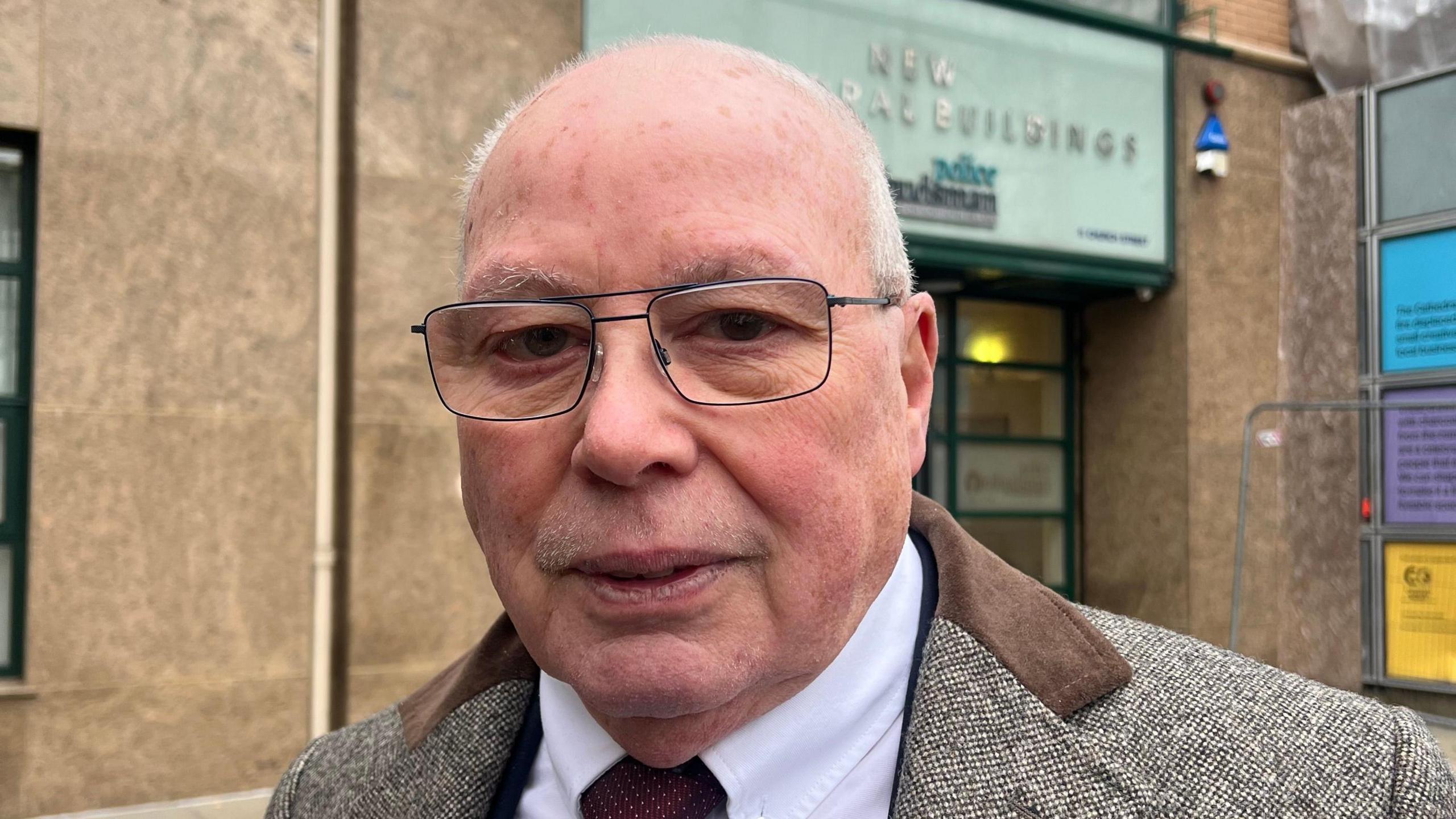 Jim Mills looking at camera. He is wearing glasses, a tweed jacket with brown suede lapels, a white shirt and a red tie with white dots. He is standing in front of a brown building with a glass front door.