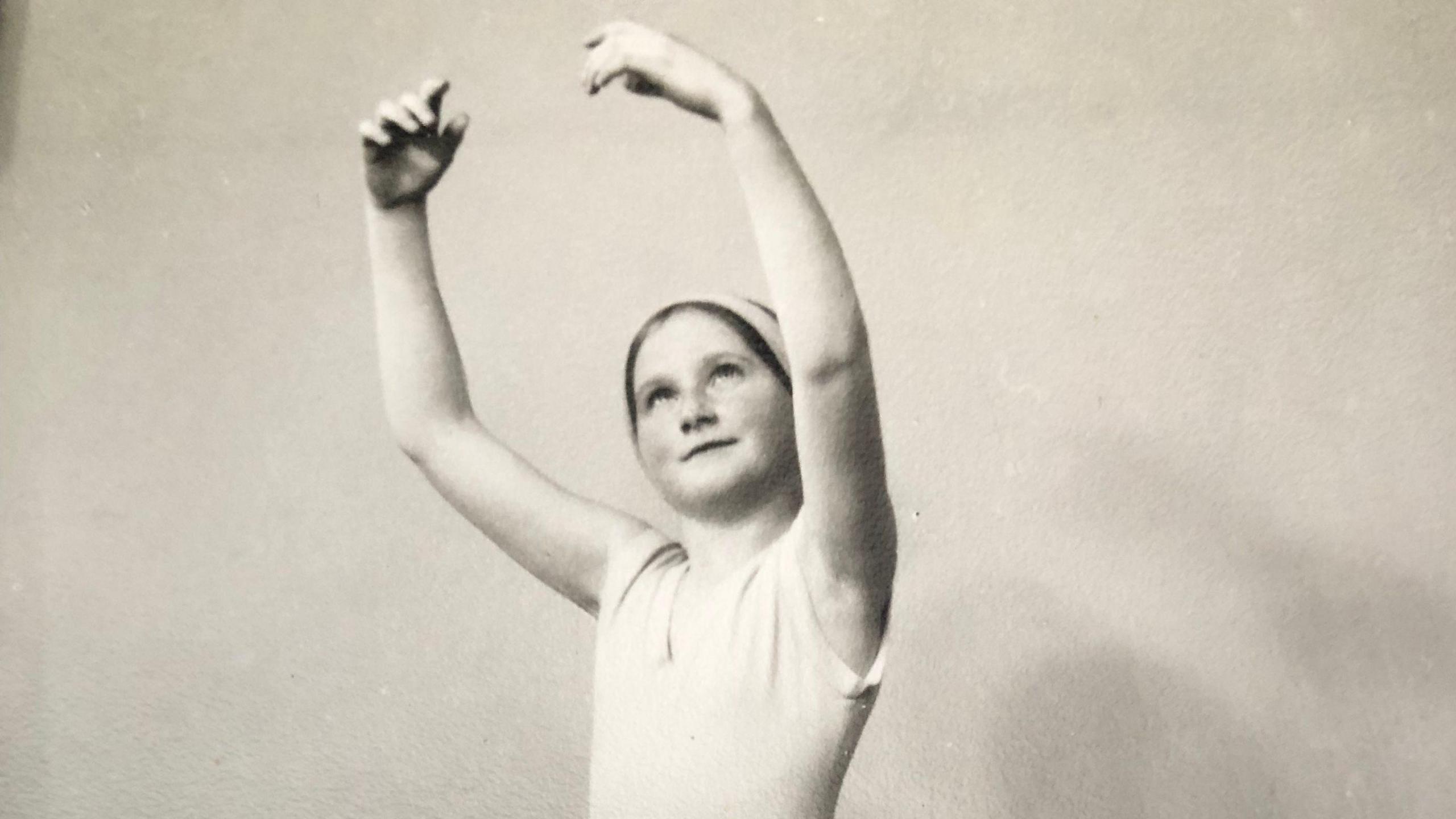 A black and white image of Helen Leonard-Taylor, aged 11, holding her hands above her head in a ballet pose and looking up slightly at her left hand. She is wearing a white leotard. 