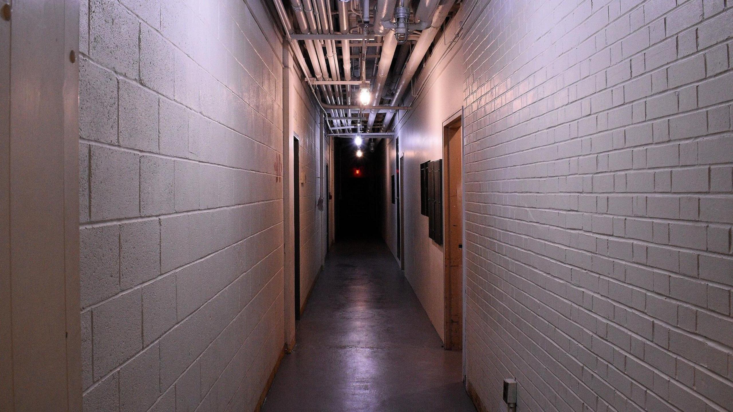 A long industrial hallway, with the walls made out of concrete blocks painted white and a cement floor. Pipes are visible running along the ceiling. 