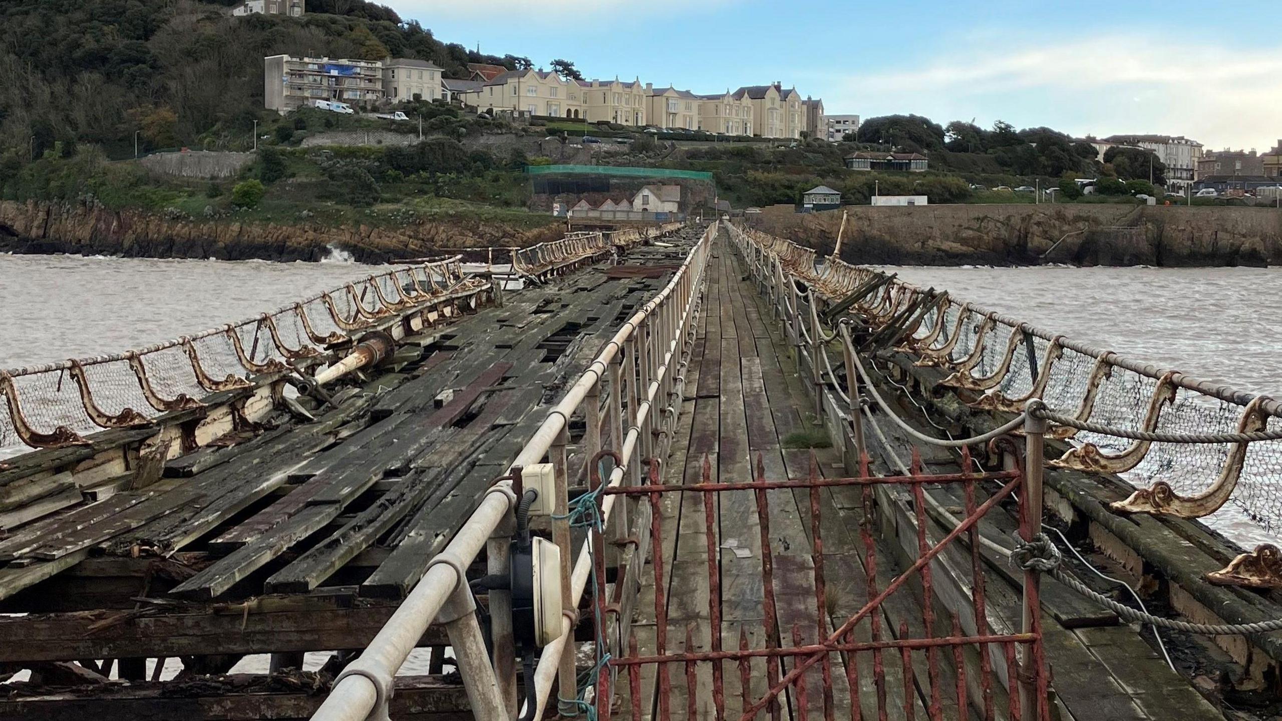 A close up image of Birnbeck Pier, taken from on the pier itself looking back towards the shore. The wooden planks look rotten and unsafe, and are lifting up from the structure.